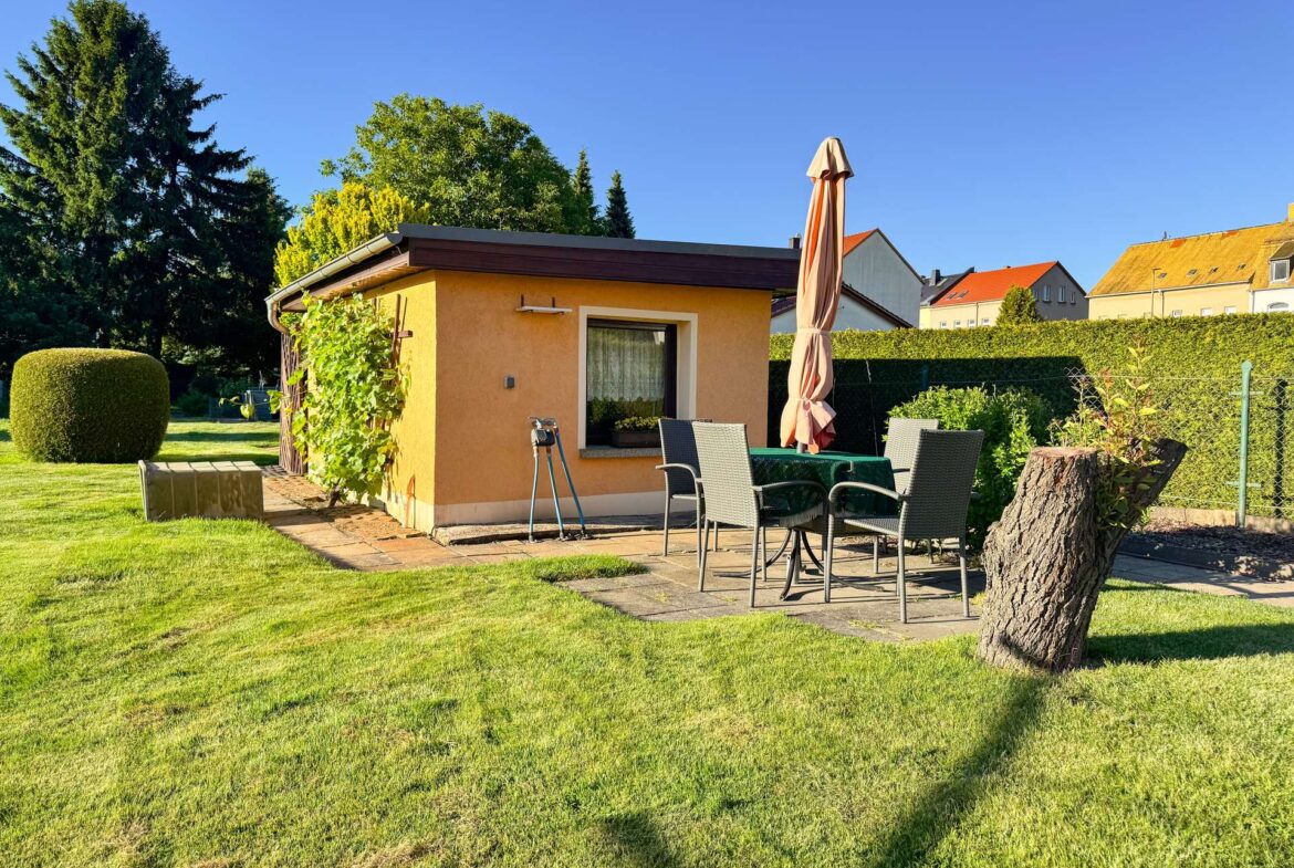 Backyard patio with orange stucco shed, beige umbrella, and gray wicker table and chairs on a stone slab.