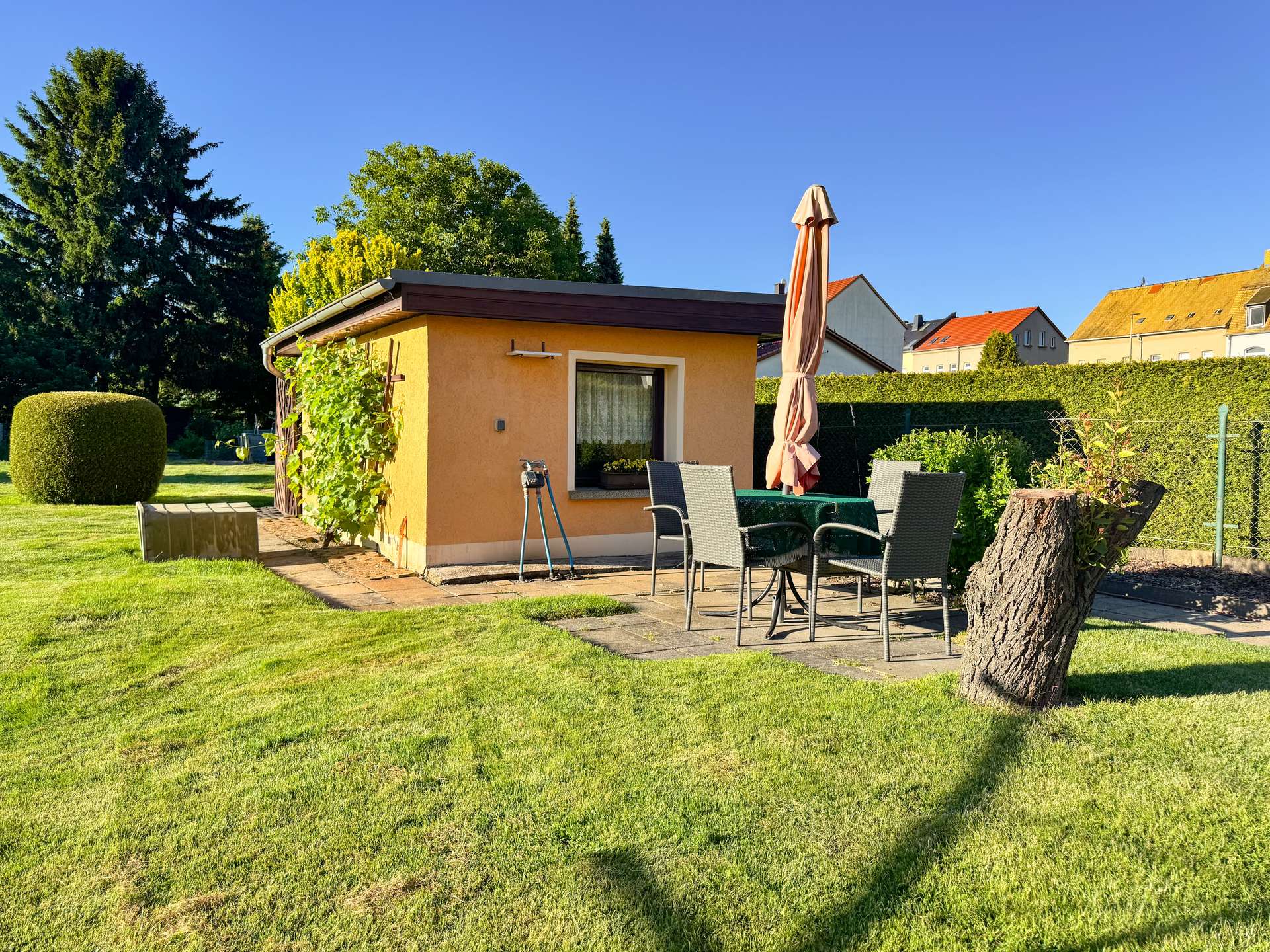 Backyard patio with orange stucco shed, beige umbrella, and gray wicker table and chairs on a stone slab.