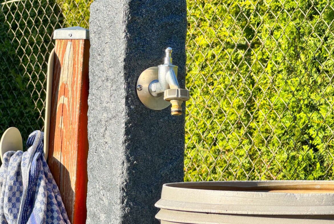 Outdoor utility area showing a silver hose bib attached to a concrete post, with a wooden post nearby and a chain-link fence in the background.