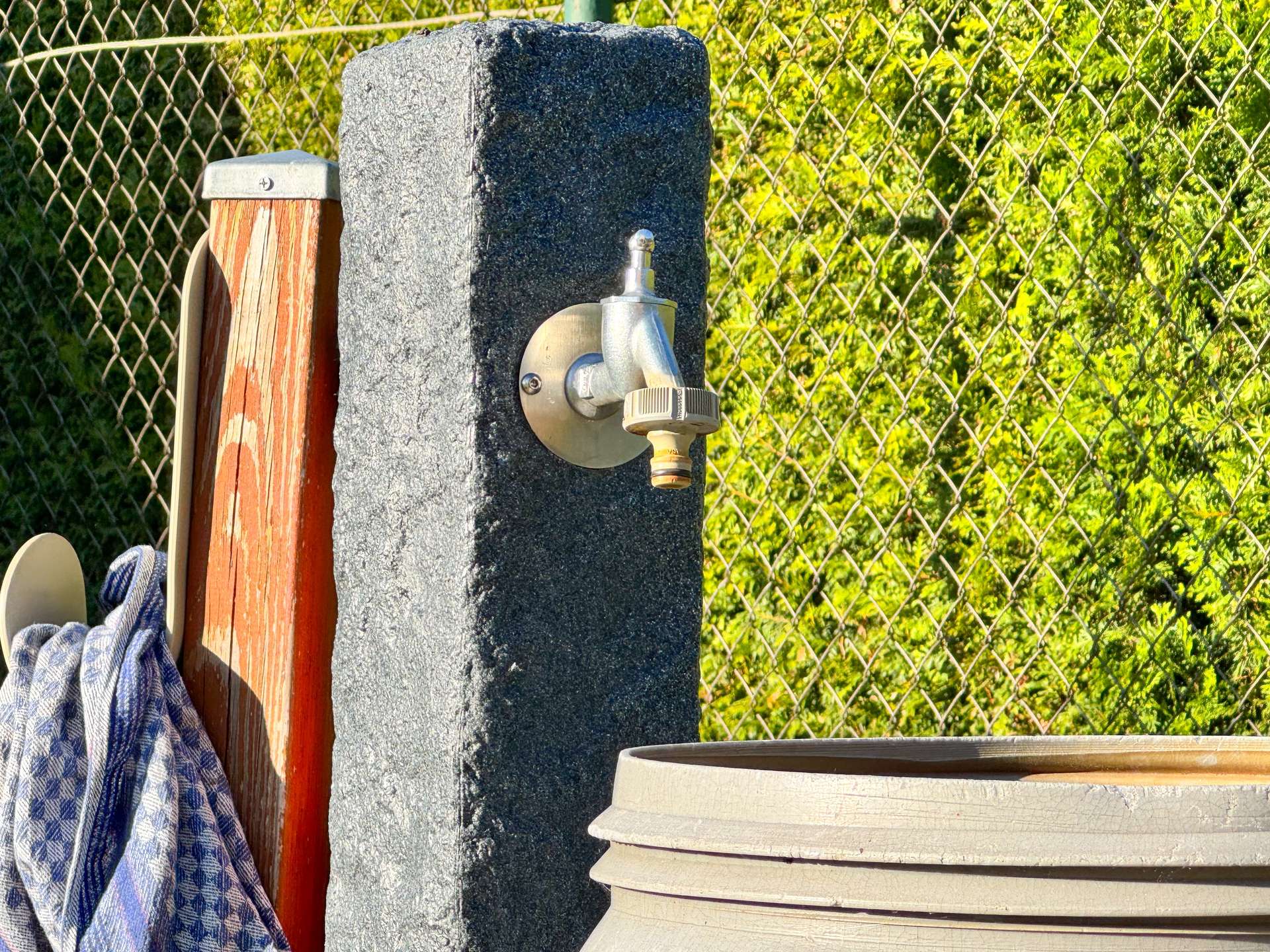 Outdoor utility area showing a silver hose bib attached to a concrete post, with a wooden post nearby and a chain-link fence in the background.