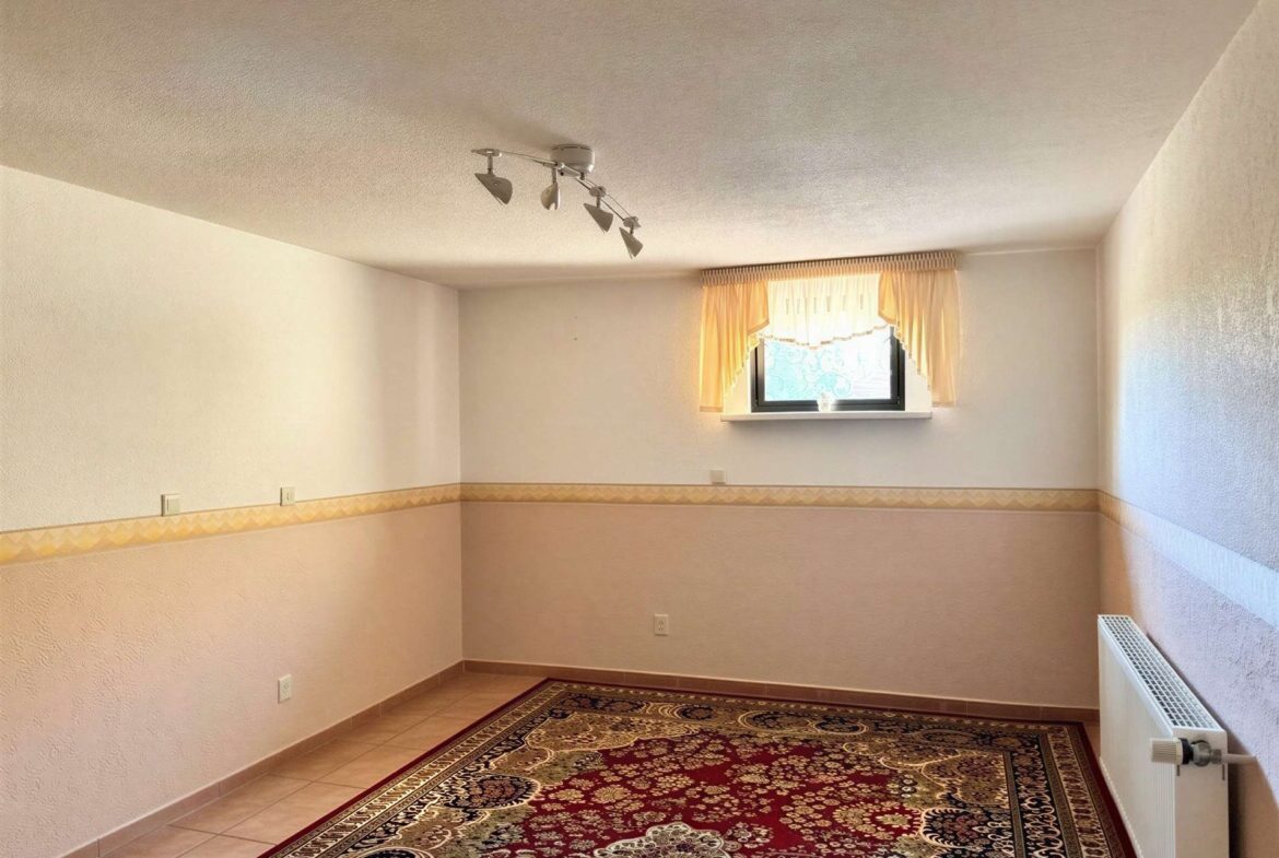 Empty room with ornate red-and-beige rug, pale walls, a small window with yellow curtains, and a radiator on the right side