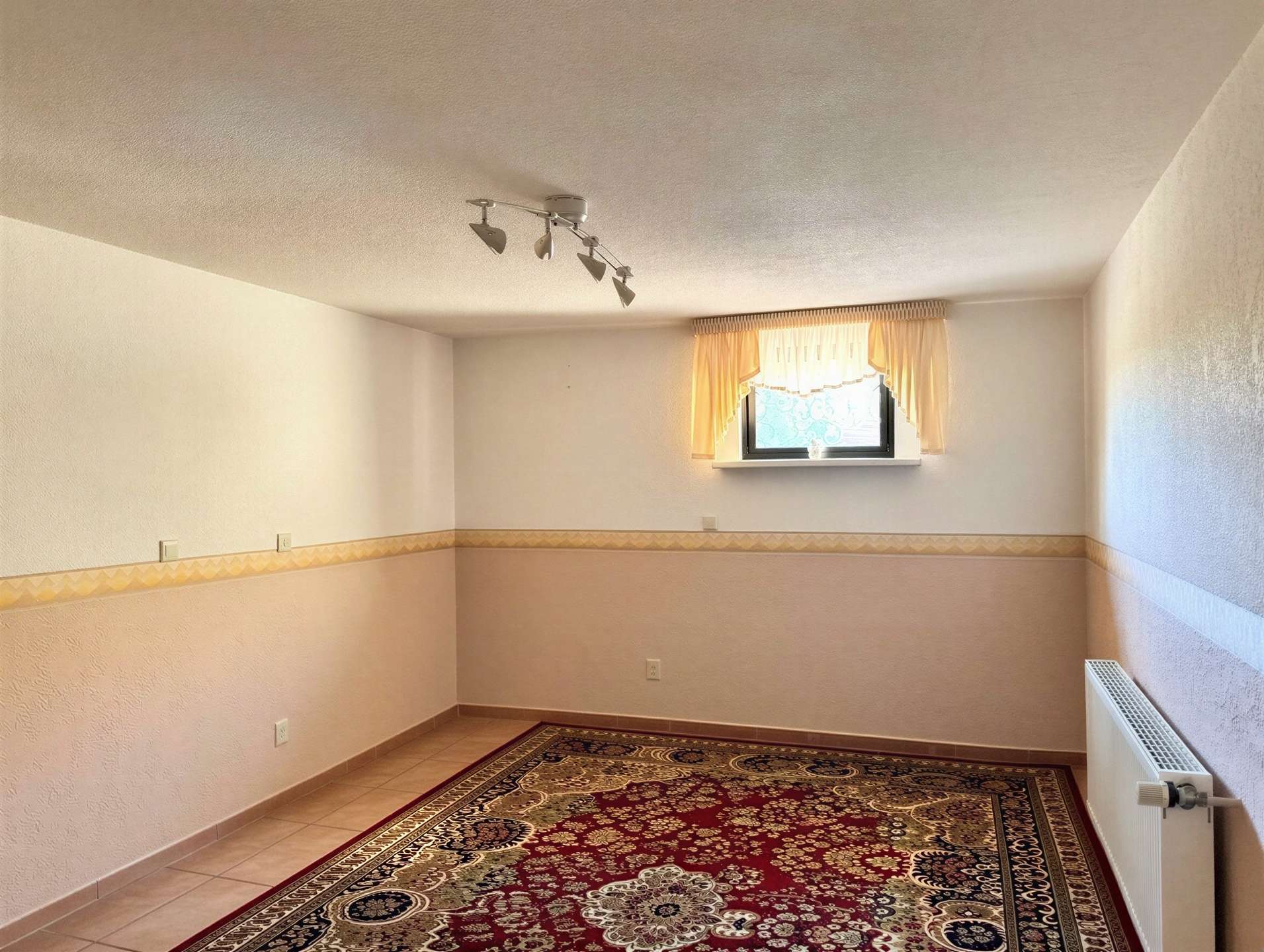 Empty room with ornate red-and-beige rug, pale walls, a small window with yellow curtains, and a radiator on the right side