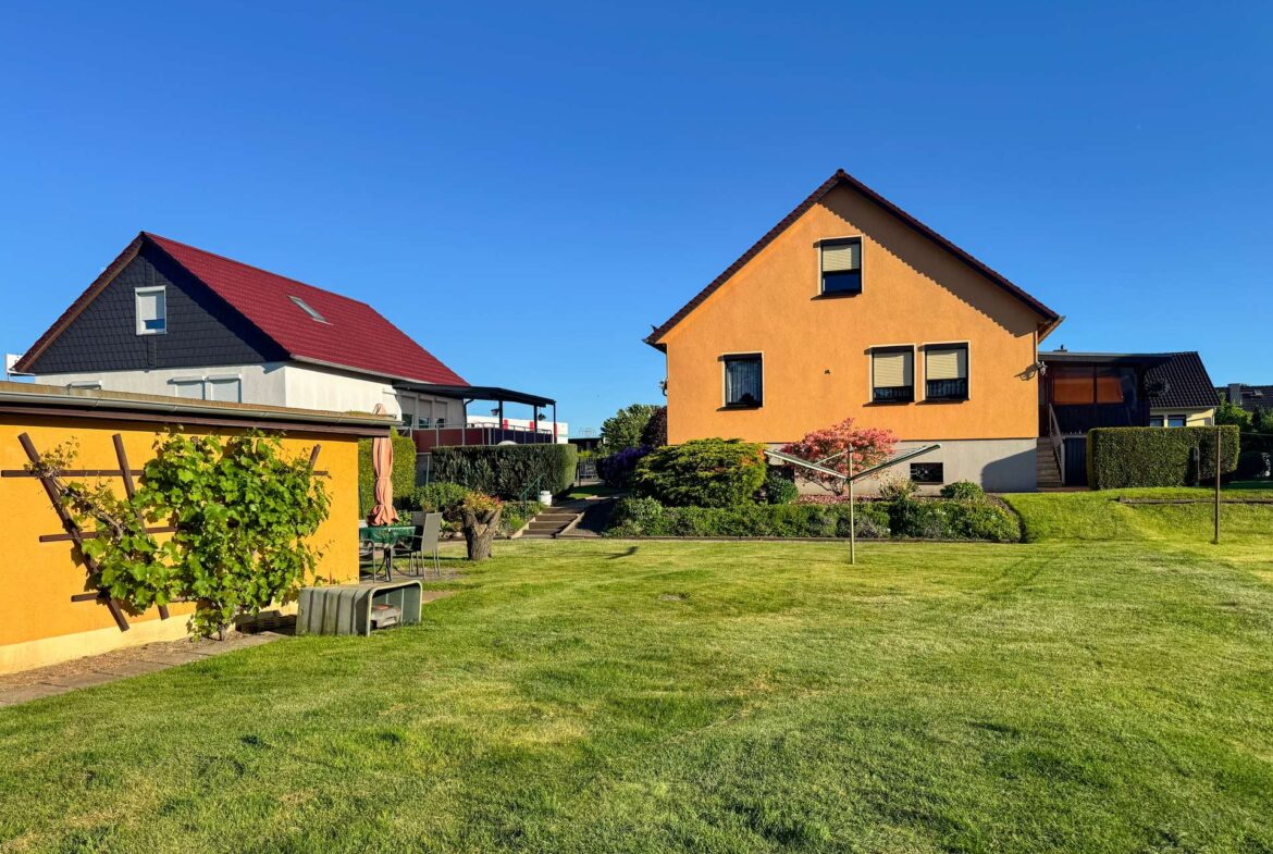 Orange two-story house with a steep gable roof, two ground-floor windows, and small upper window, set in a landscaped yard under a bright blue sky.