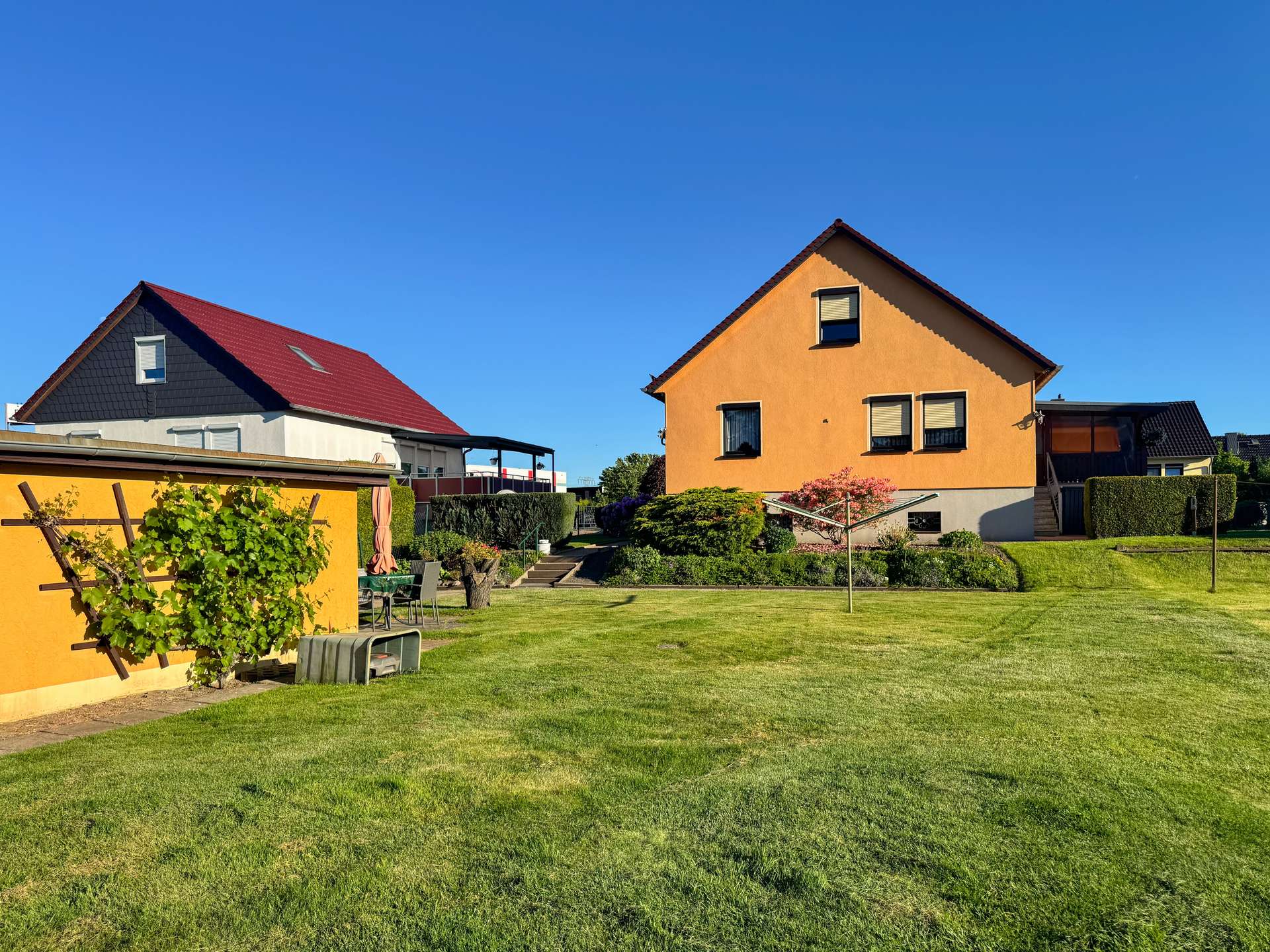 Orange two-story house with a steep gable roof, two ground-floor windows, and small upper window, set in a landscaped yard under a bright blue sky.