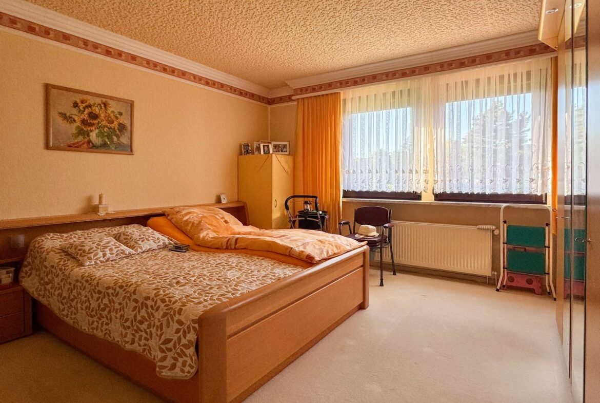 Two wooden beds with patterned bedding in a warm beige bedroom, sunflowers painting on the wall, and large windows with sheer curtains and orange drapes.