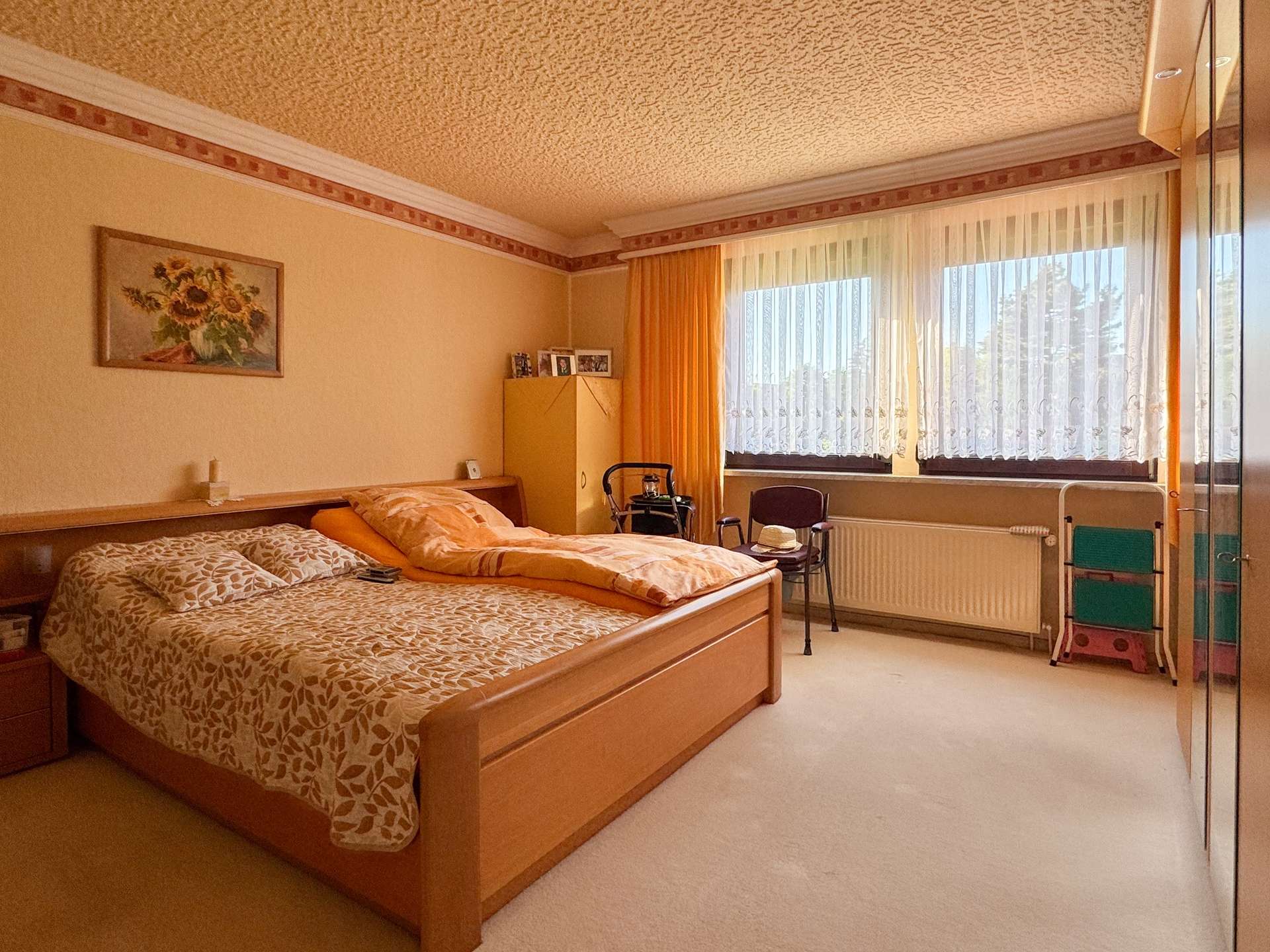 Two wooden beds with patterned bedding in a warm beige bedroom, sunflowers painting on the wall, and large windows with sheer curtains and orange drapes.