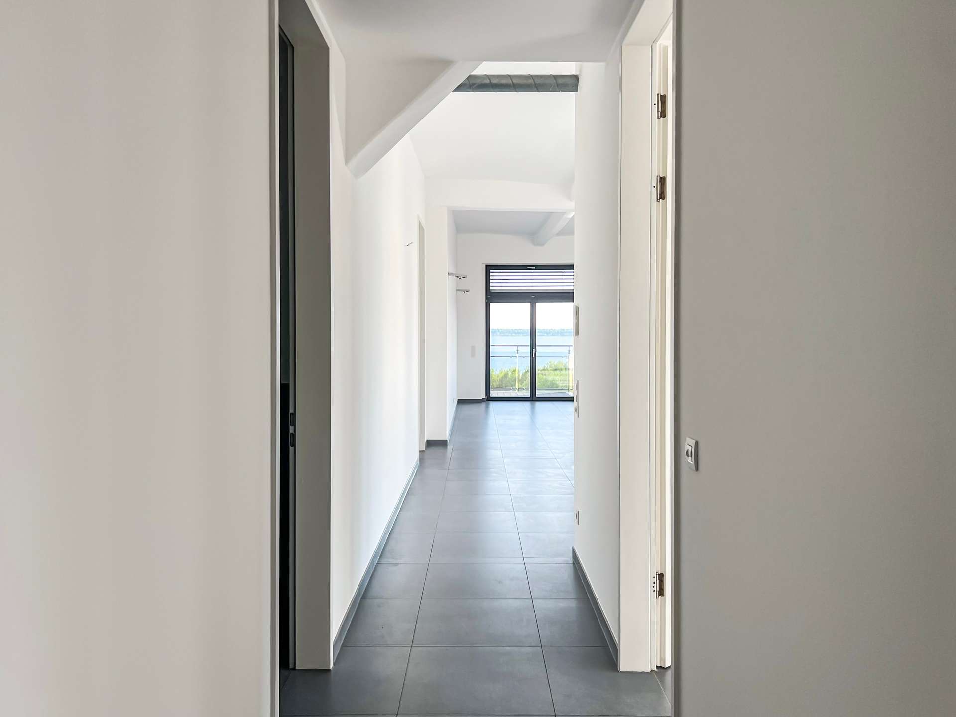 Long indoor hallway leading to a glass door with a balcony and ocean view beyond.