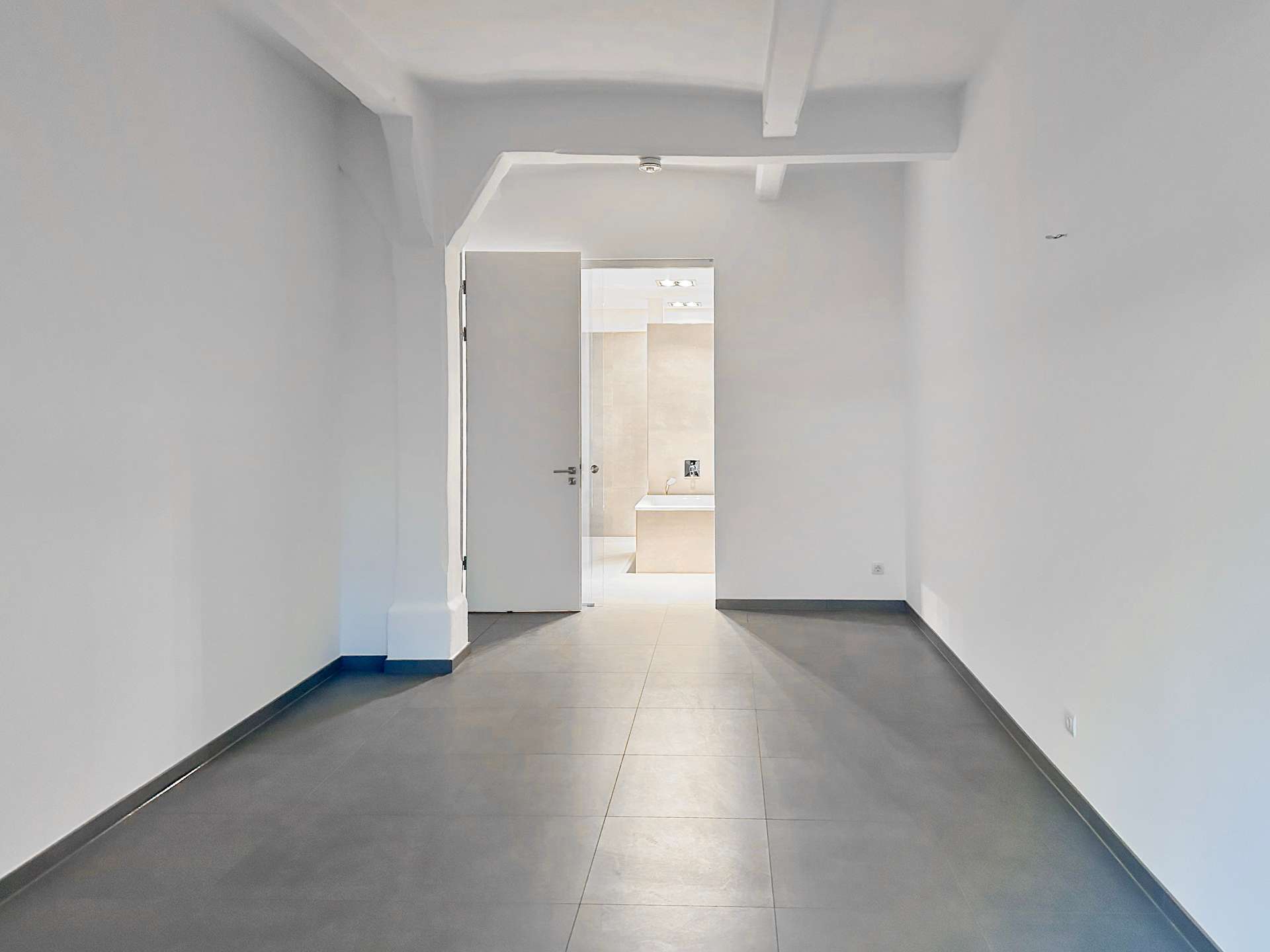 Empty gray-tiled hallway with white walls and an open door at the far end revealing a bright room beyond.
