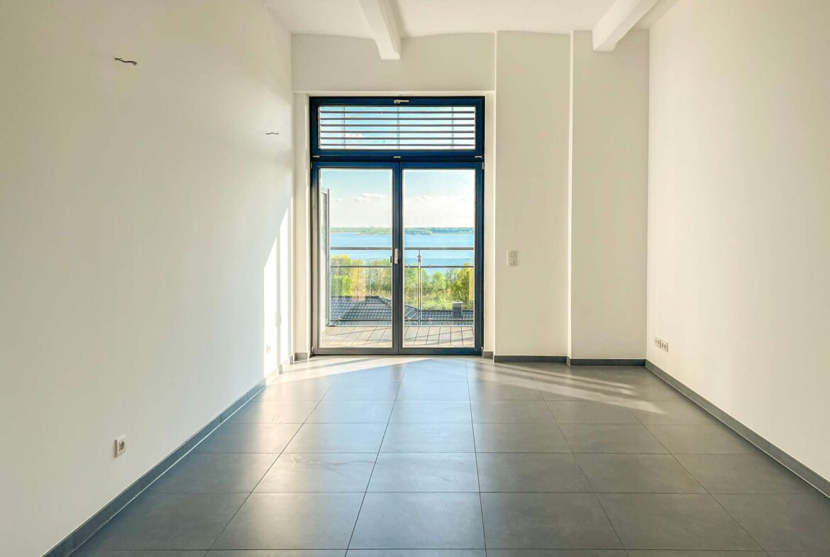 Bright empty hallway with a glass door opening to a balcony and a lake view beyond. The space has light walls and large gray tiled floor.