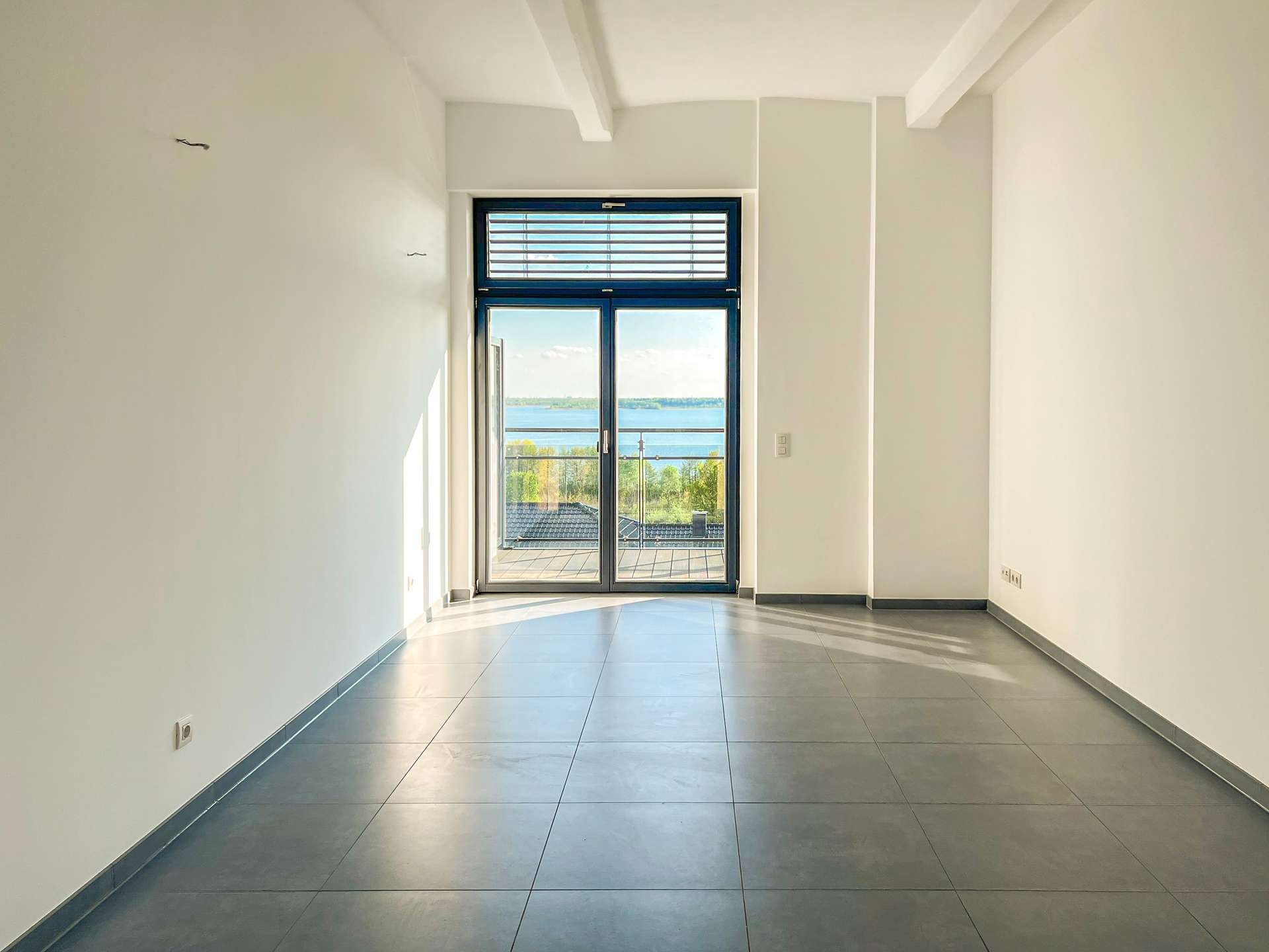 Bright empty hallway with a glass door opening to a balcony and a lake view beyond. The space has light walls and large gray tiled floor.