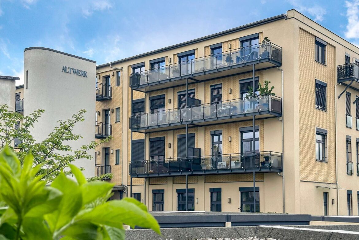 Modern beige apartment building with glass-balcony railings under a blue sky; foreground plants frame the scene.