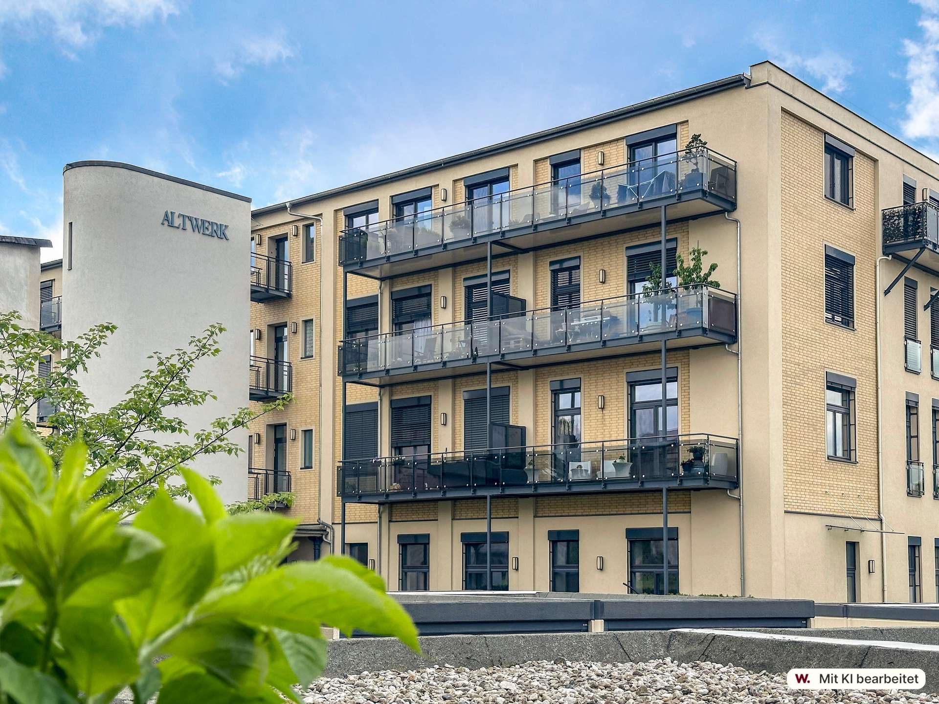 Modern beige apartment building with glass-balcony railings under a blue sky; foreground plants frame the scene.