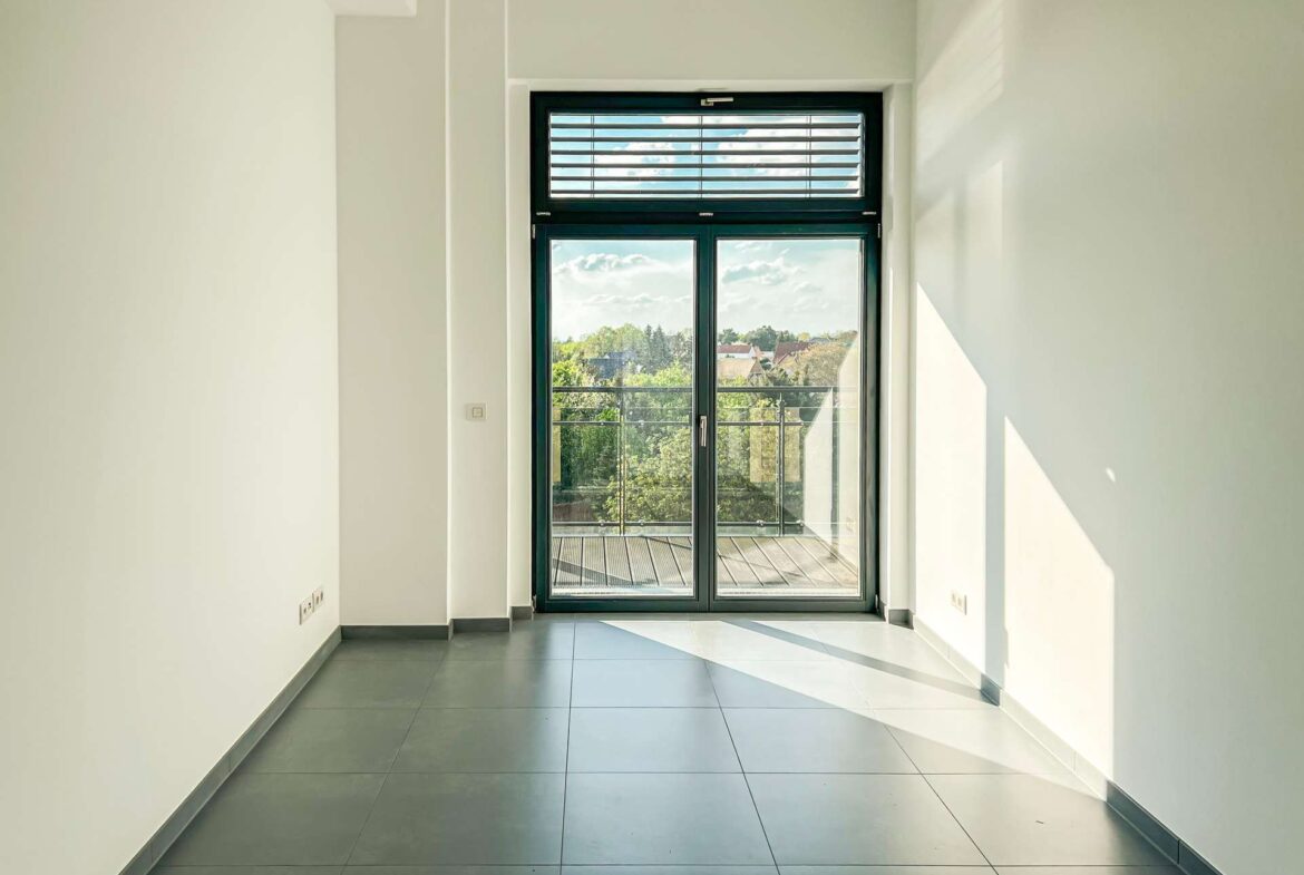 Bright, empty hallway leading to a large glass door with a balcony and views of trees and rooftops outside.