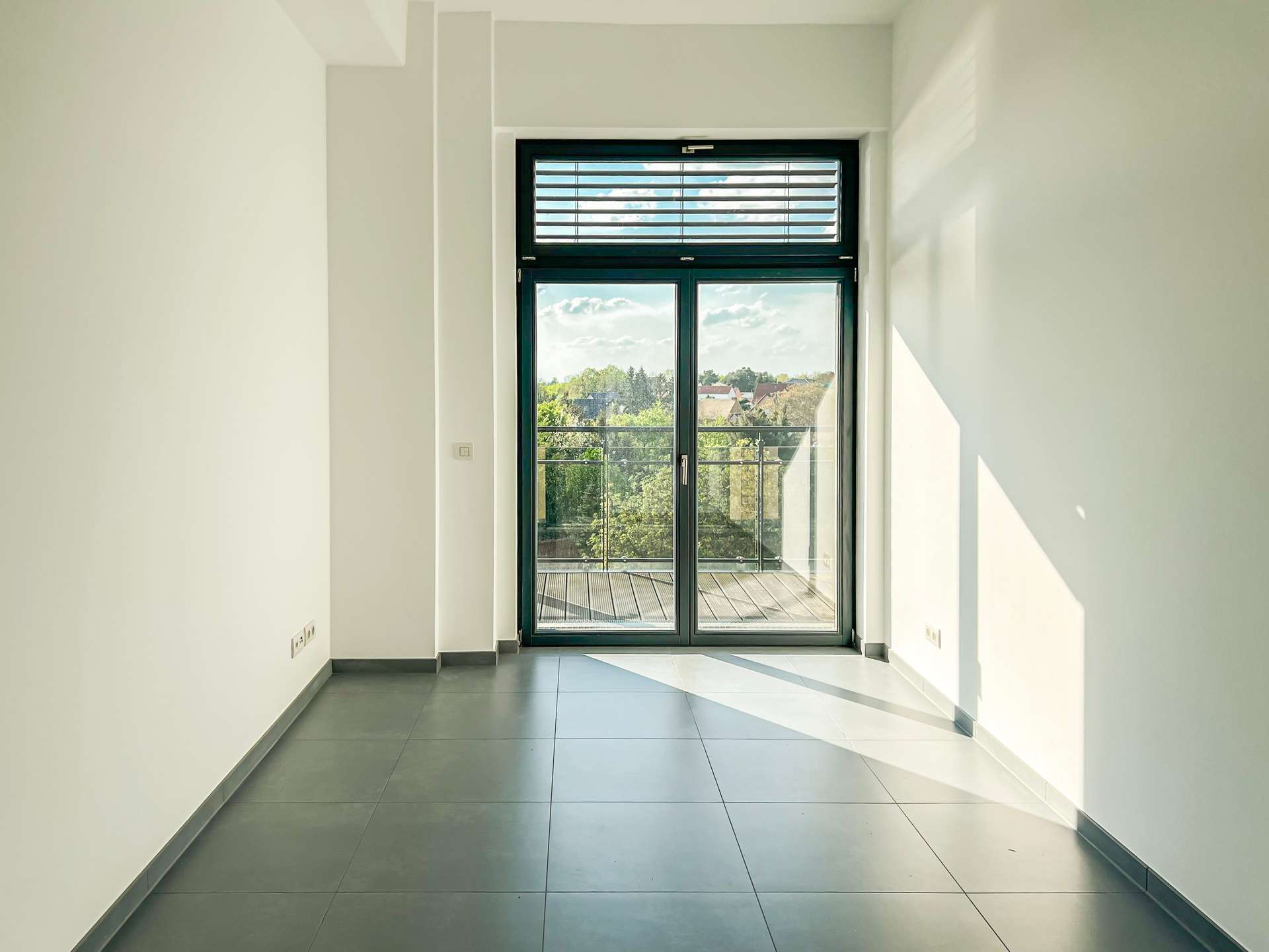 Bright, empty hallway leading to a large glass door with a balcony and views of trees and rooftops outside.