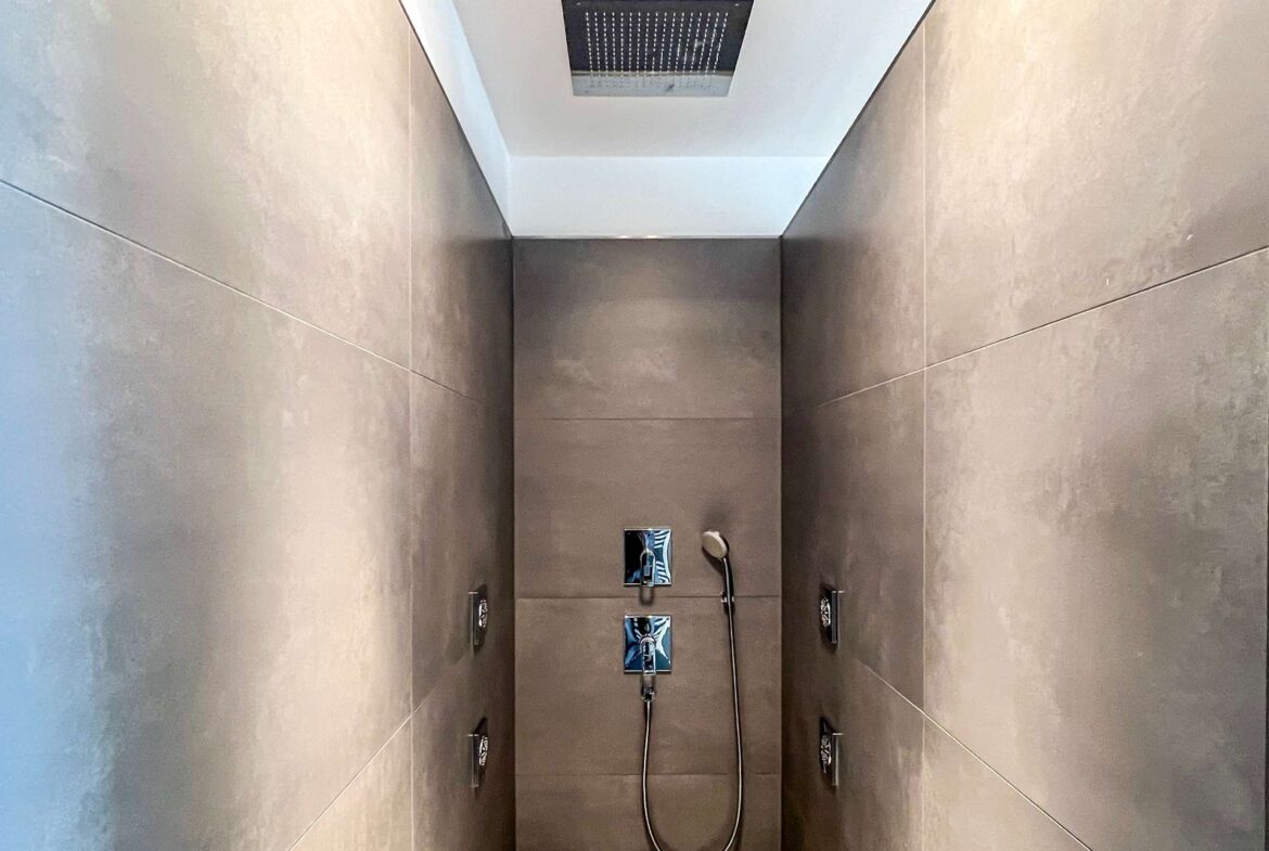 Shower enclosure with a square ceiling rainfall shower and a handheld shower on the wall, covered in dark gray tiles.