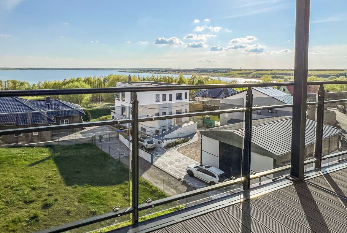 Balcony railing with a view of houses, a parking area, a grassy yard, and a large lake beyond under a bright blue sky.