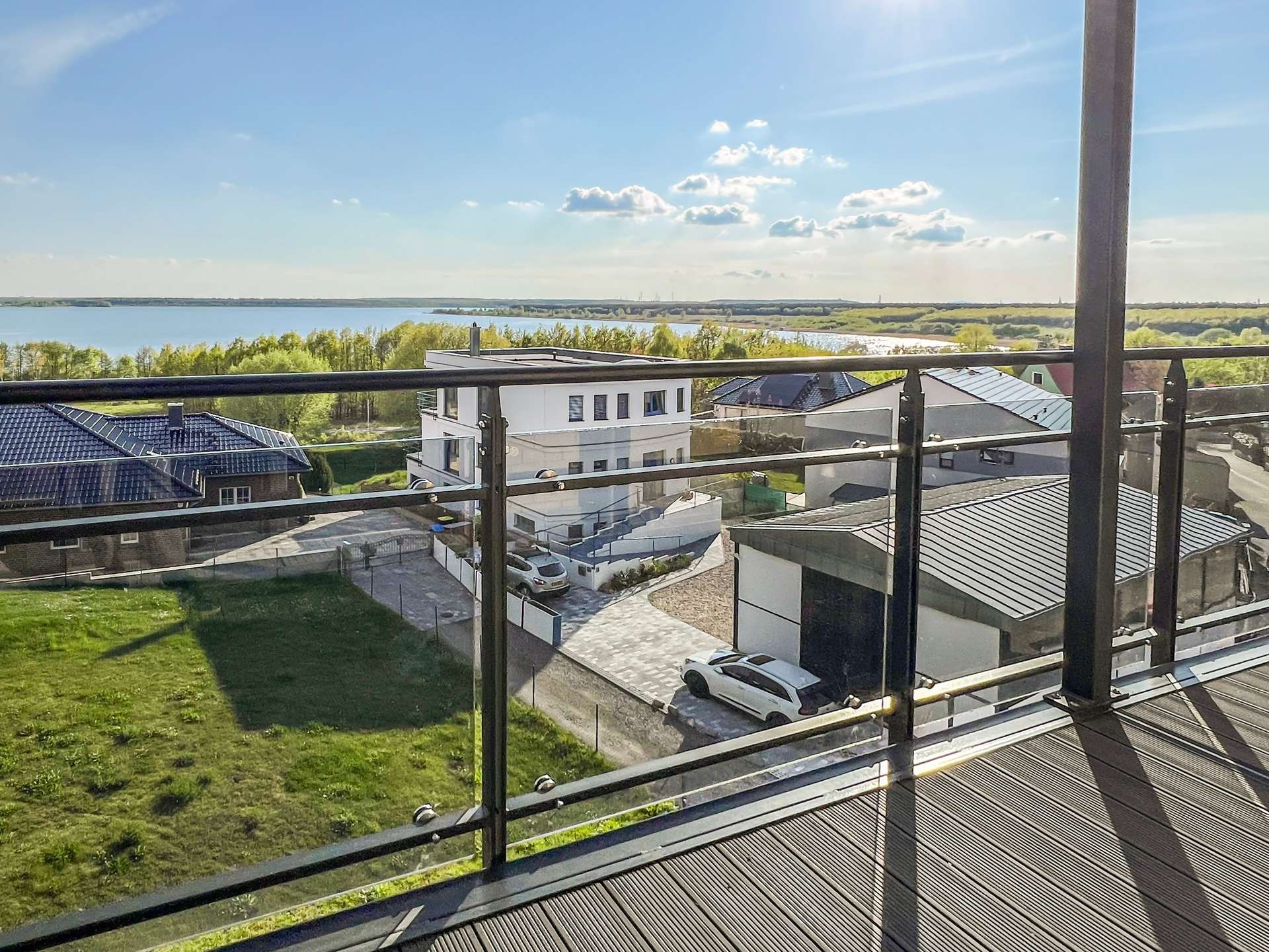 Balcony railing with a view of houses, a parking area, a grassy yard, and a large lake beyond under a bright blue sky.