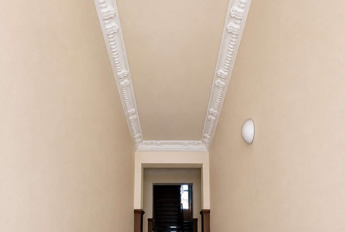 Narrow beige hallway with ornate white crown molding along the ceiling, leading to a dark wooden staircase; a round wall sconce is on the right.