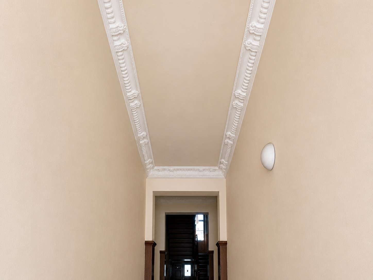 Narrow beige hallway with ornate white crown molding along the ceiling, leading to a dark wooden staircase; a round wall sconce is on the right.