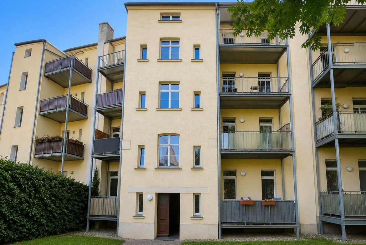 Five-story beige apartment building with gray balconies and a central entrance, under a clear blue sky and green lawn.