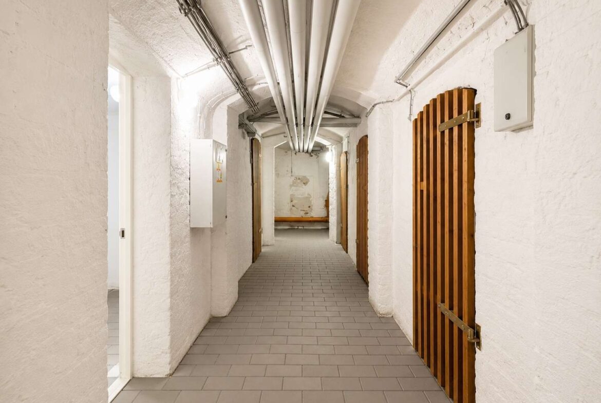 Long white-walled basement corridor with exposed pipes on the ceiling, gray tile floor, and wooden slatted doors along the right side