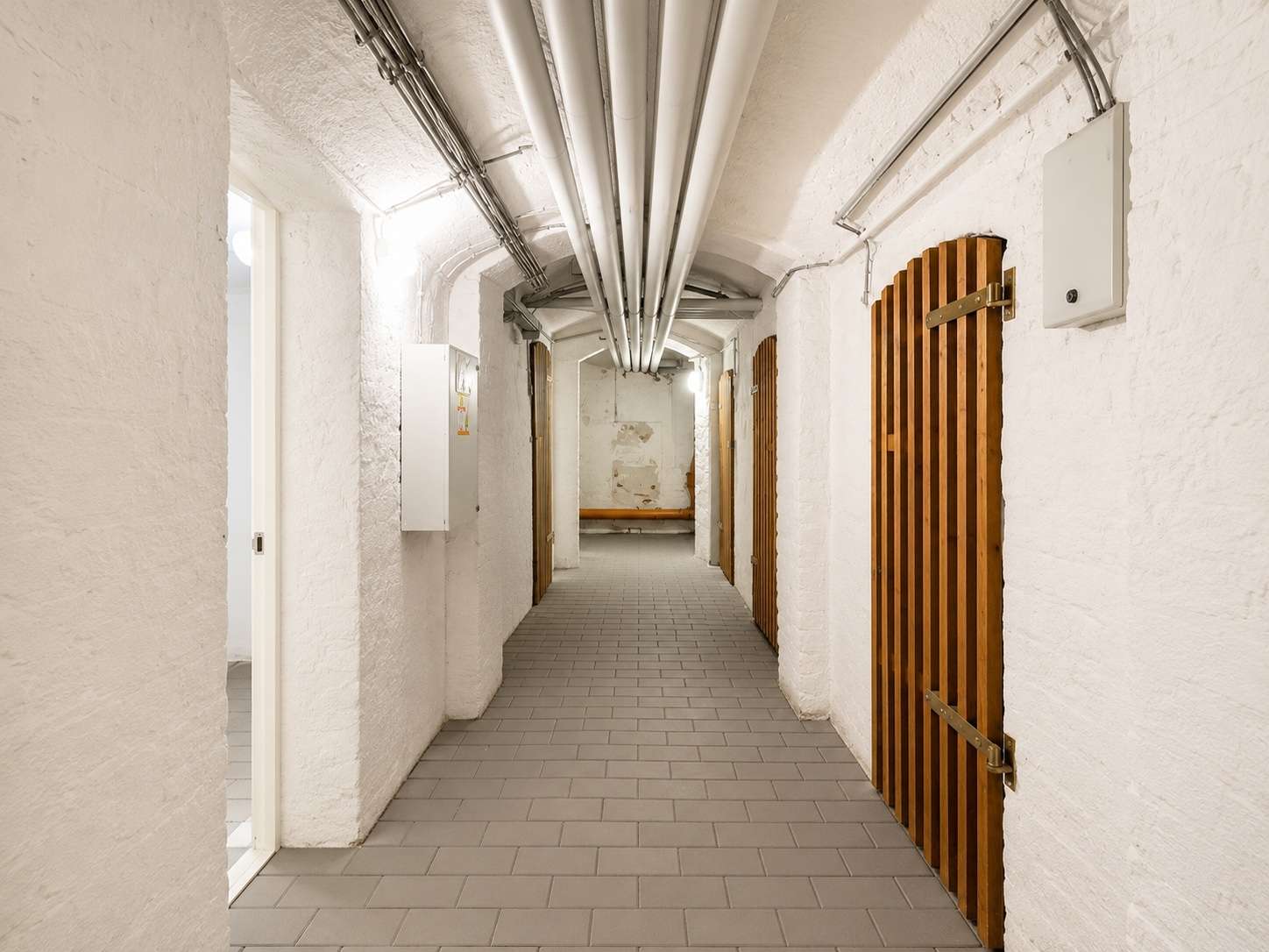 Long white-walled basement corridor with exposed pipes on the ceiling, gray tile floor, and wooden slatted doors along the right side
