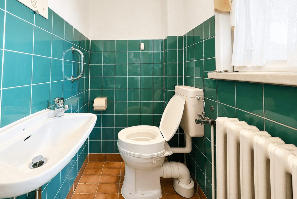 Small bathroom with teal tiled walls, a white toilet and sink, and a radiator by a window with a sheer curtain.