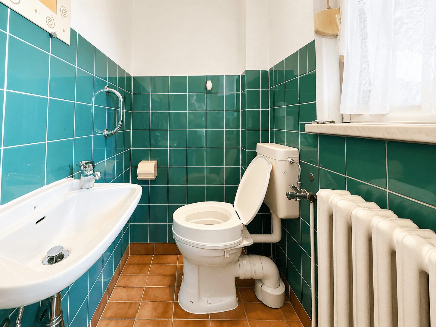 Small bathroom with teal tiled walls, a white toilet and sink, and a radiator by a window with a sheer curtain.