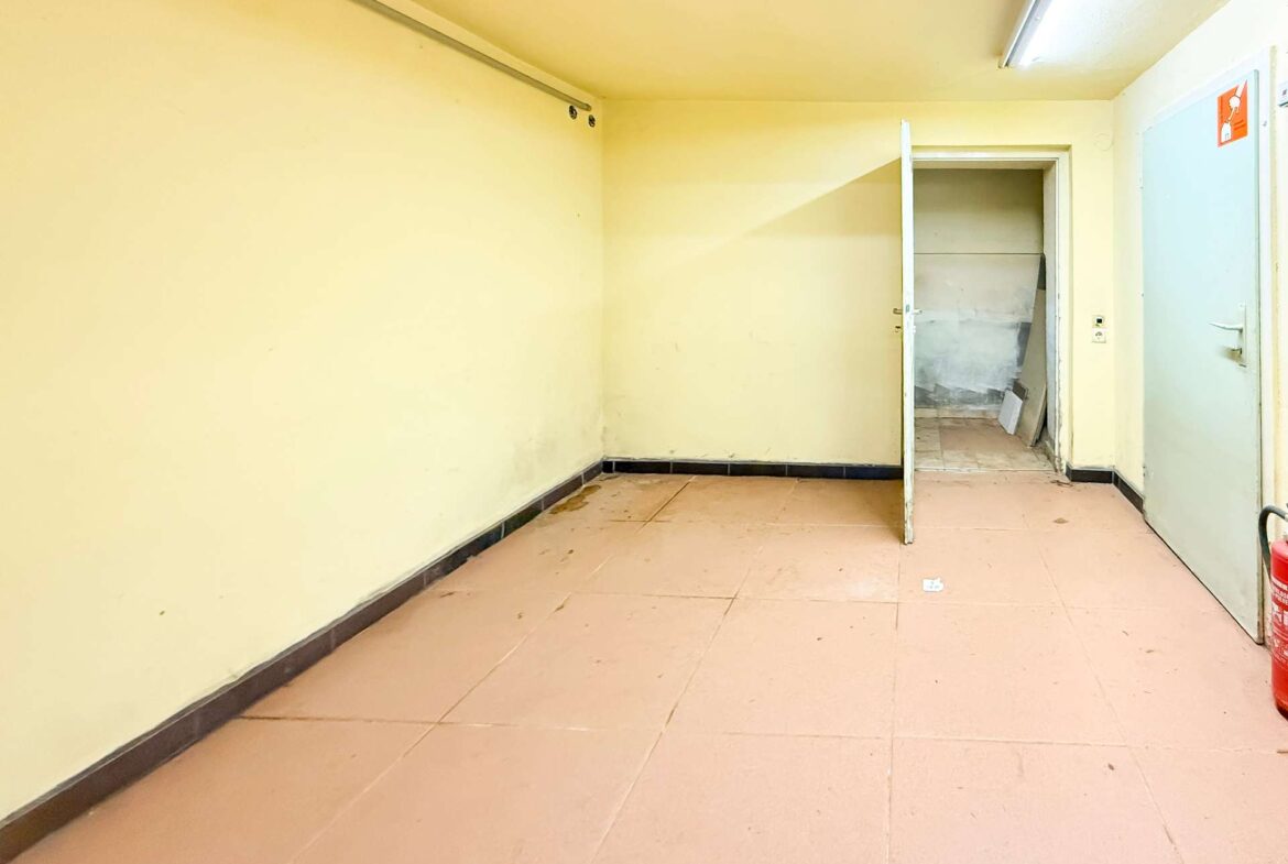 Empty yellow-walled room with pink tile floor; open doorway leads to a small storage area filled with debris, and a red fire extinguisher sits in the right corner.