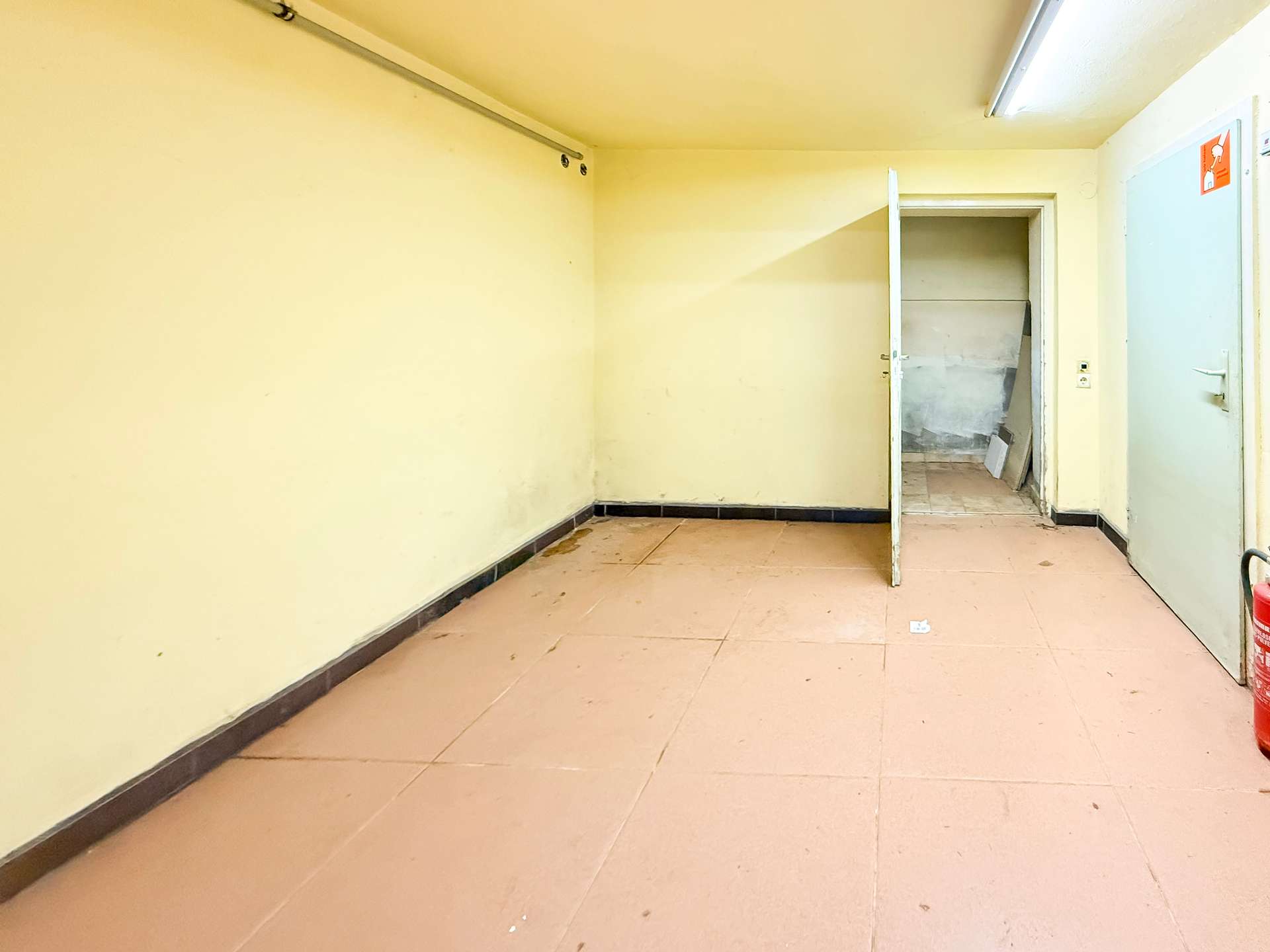 Empty yellow-walled room with pink tile floor; open doorway leads to a small storage area filled with debris, and a red fire extinguisher sits in the right corner.