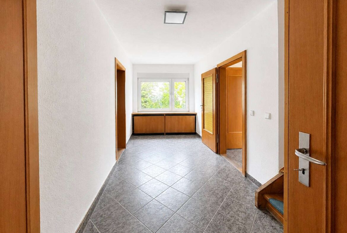 Bright, empty hallway with white walls, gray tiled floor, and wooden doors, ending at a window with a low wooden cabinet beneath it.