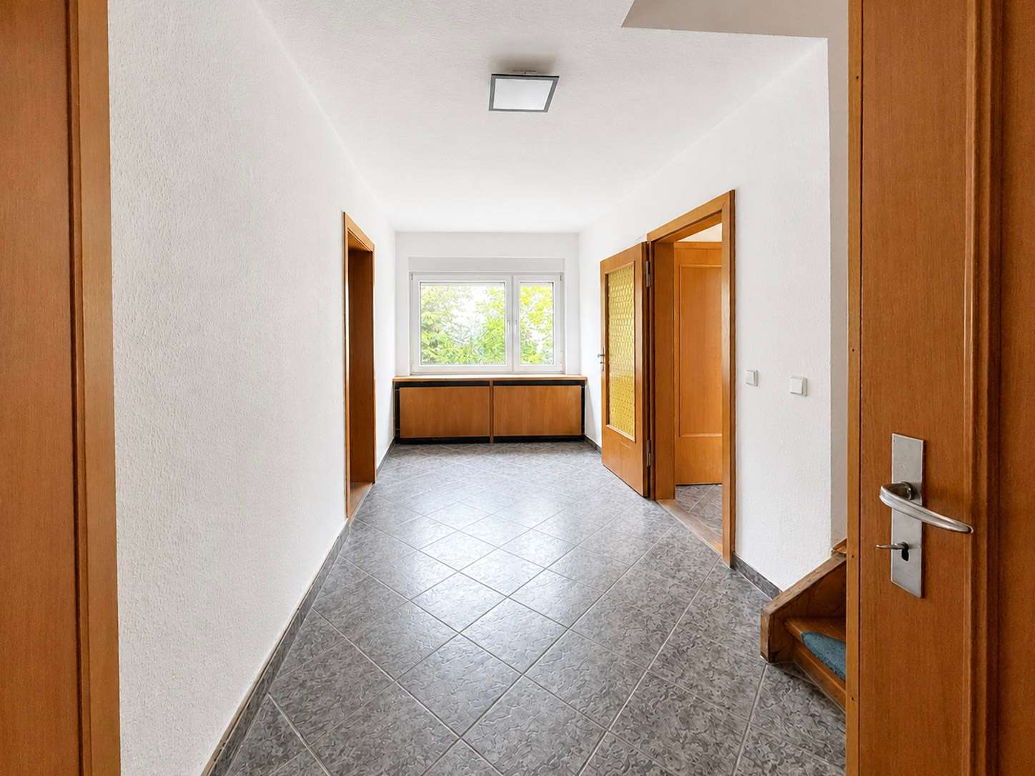 Bright, empty hallway with white walls, gray tiled floor, and wooden doors, ending at a window with a low wooden cabinet beneath it.