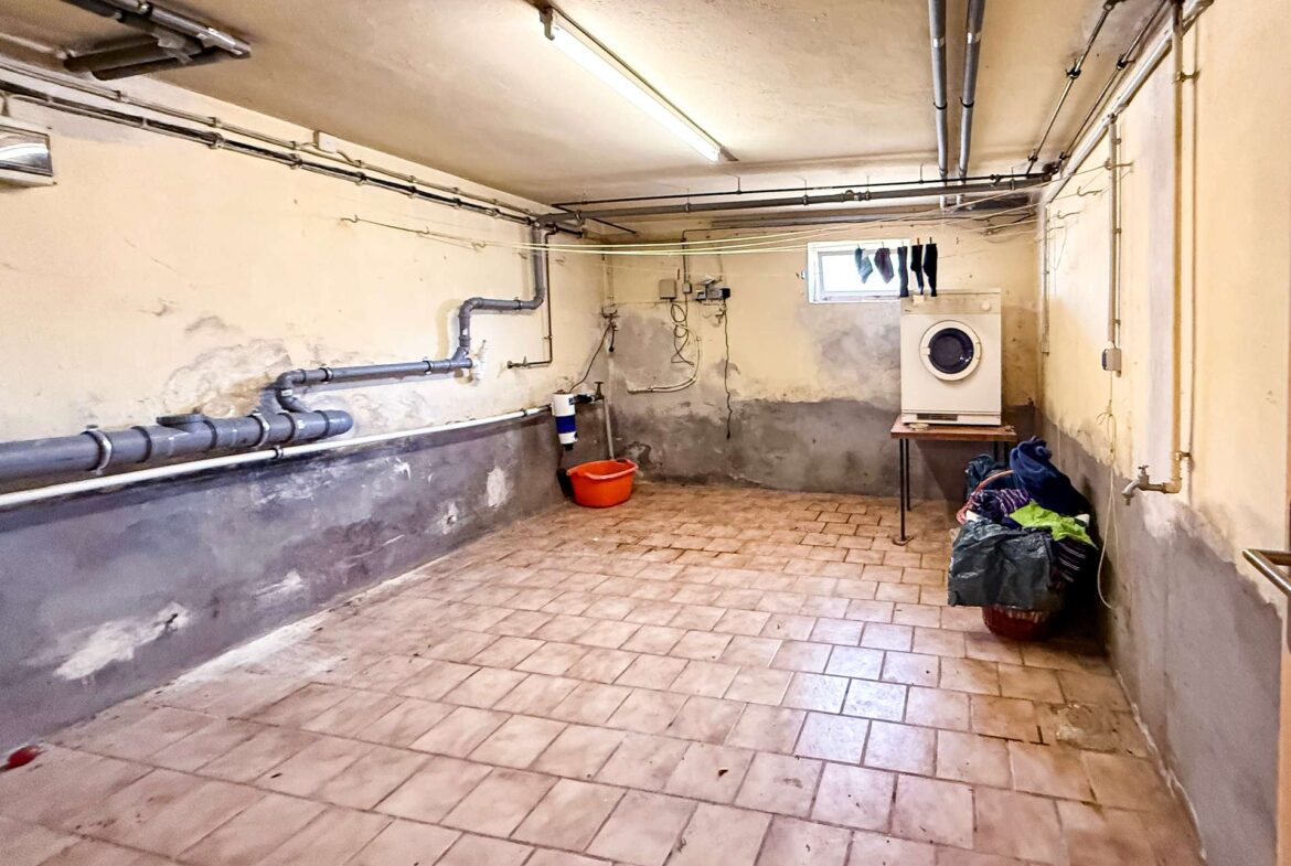 Basement laundry room with exposed pipes, washing machine on a wooden stand, clothesline with socks, and a bucket on the tiled floor