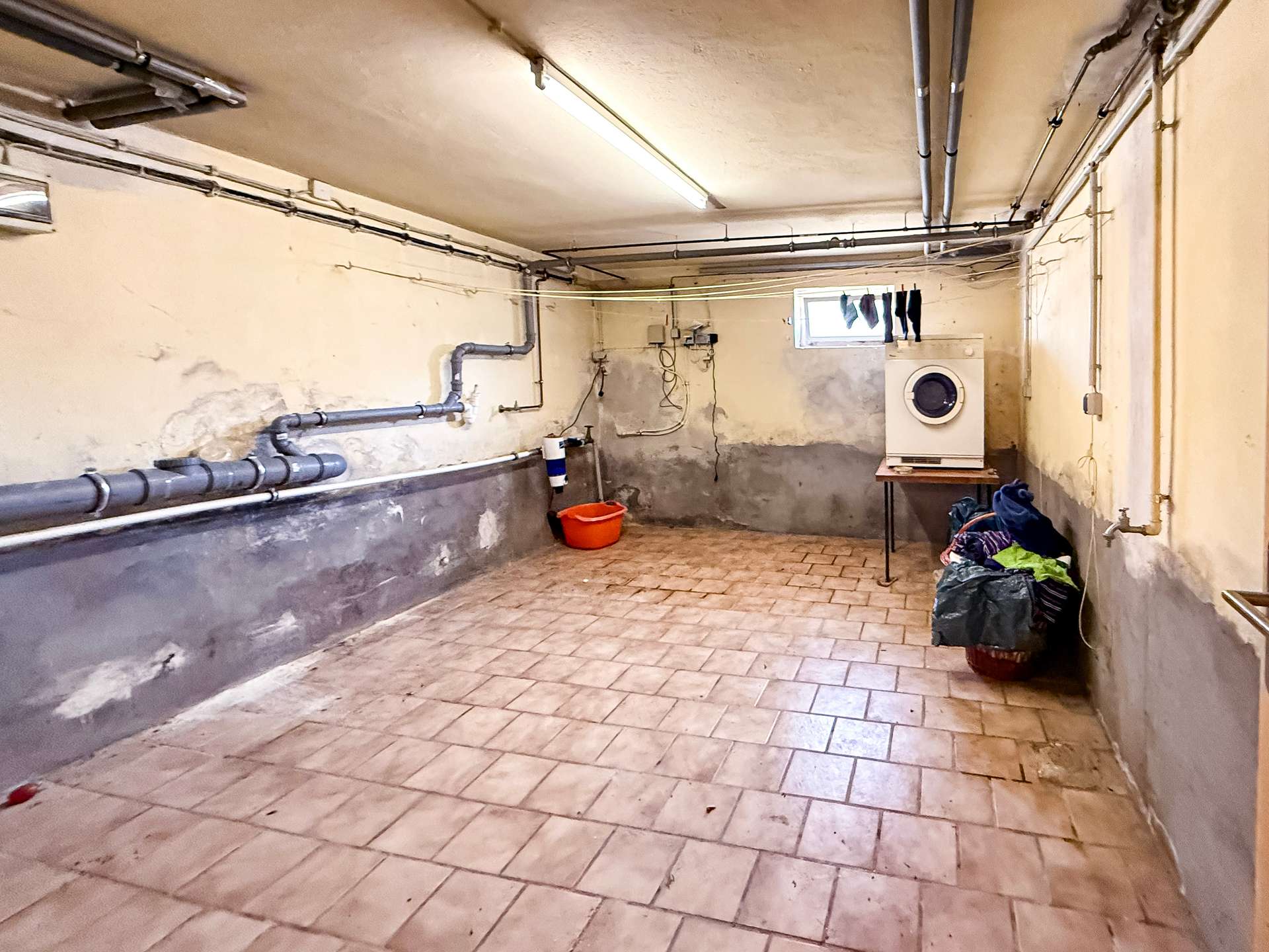 Basement laundry room with exposed pipes, washing machine on a wooden stand, clothesline with socks, and a bucket on the tiled floor