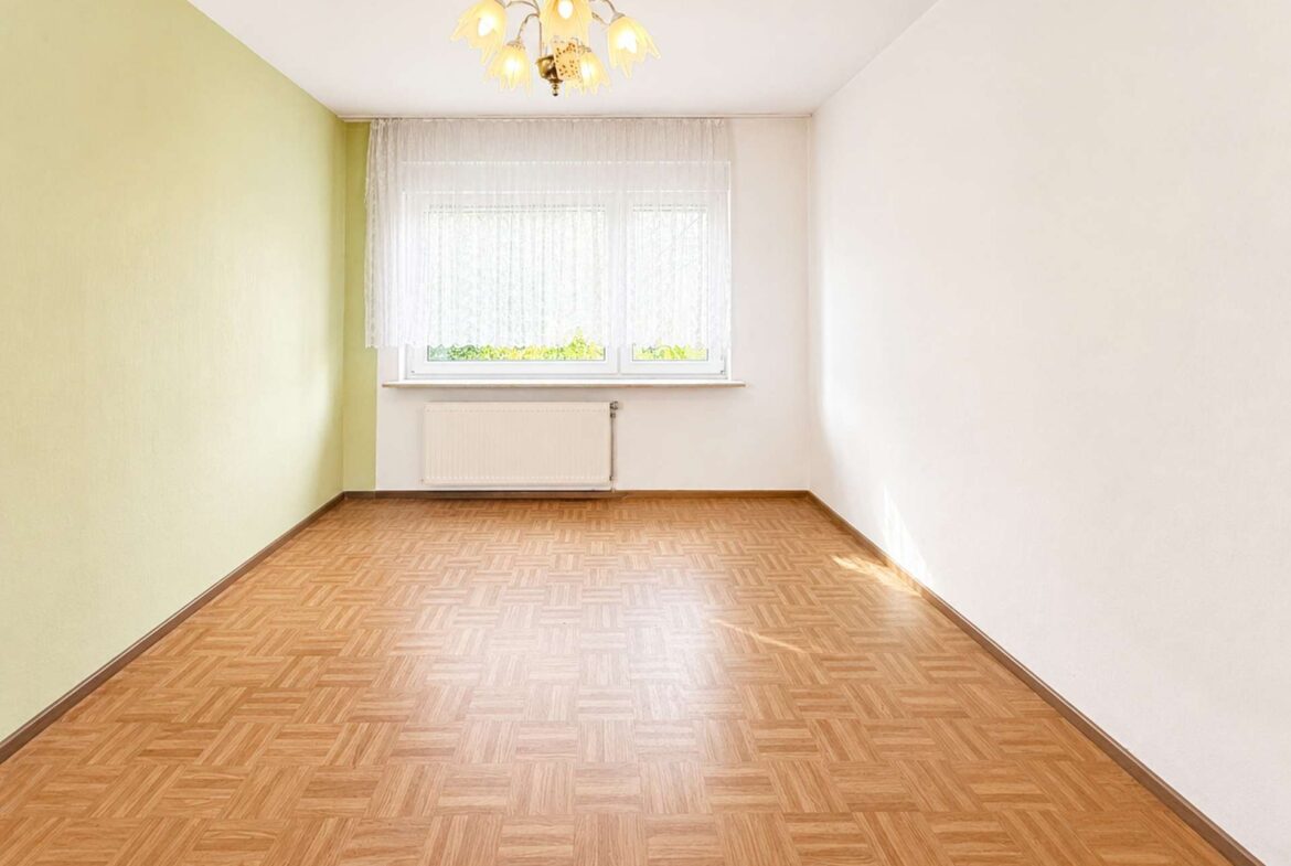 Empty rectangular room with a green left wall, white walls, a window with lace curtains, radiator beneath, and parquet floor.