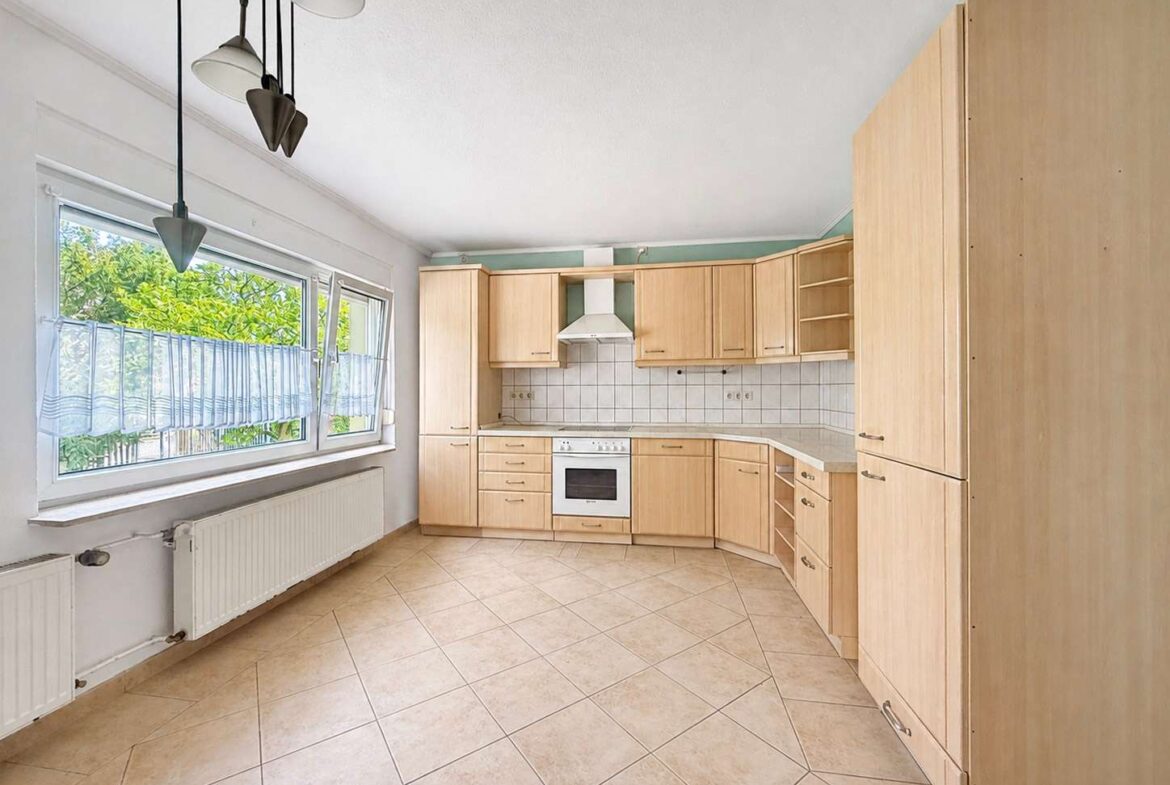 Bright L-shaped kitchen with light wood cabinets, beige tiled floor, white tile backsplash, and a white range hood over a stove. Large window with sheer blinds and a radiator along the wall.