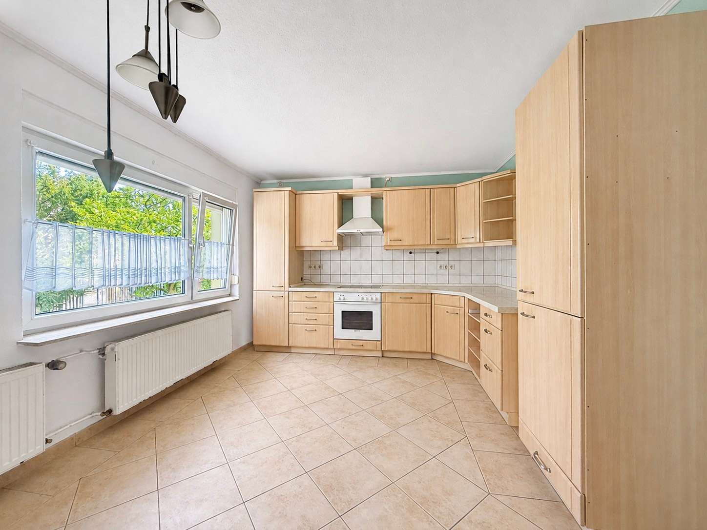 Bright L-shaped kitchen with light wood cabinets, beige tiled floor, white tile backsplash, and a white range hood over a stove. Large window with sheer blinds and a radiator along the wall.
