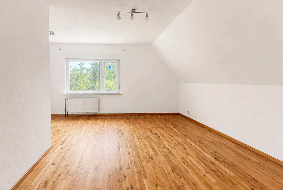 Bright, empty attic room with white walls, a sloped ceiling on the right, and a wooden floor under a small window with a radiator beneath it