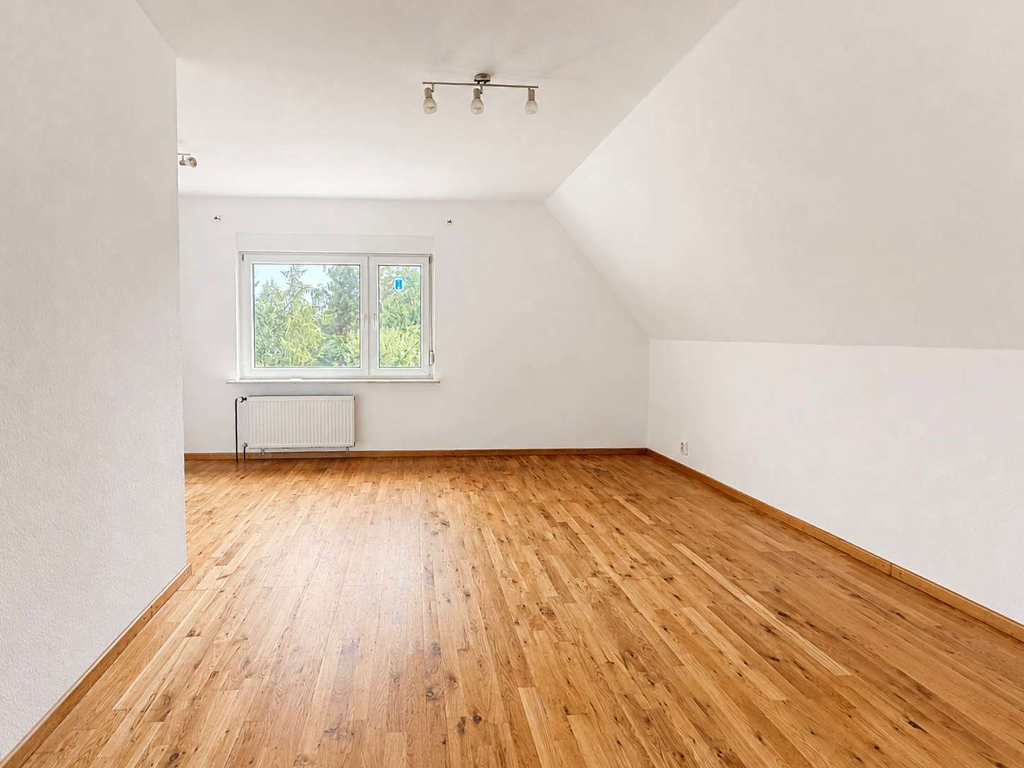 Bright, empty attic room with white walls, a sloped ceiling on the right, and a wooden floor under a small window with a radiator beneath it
