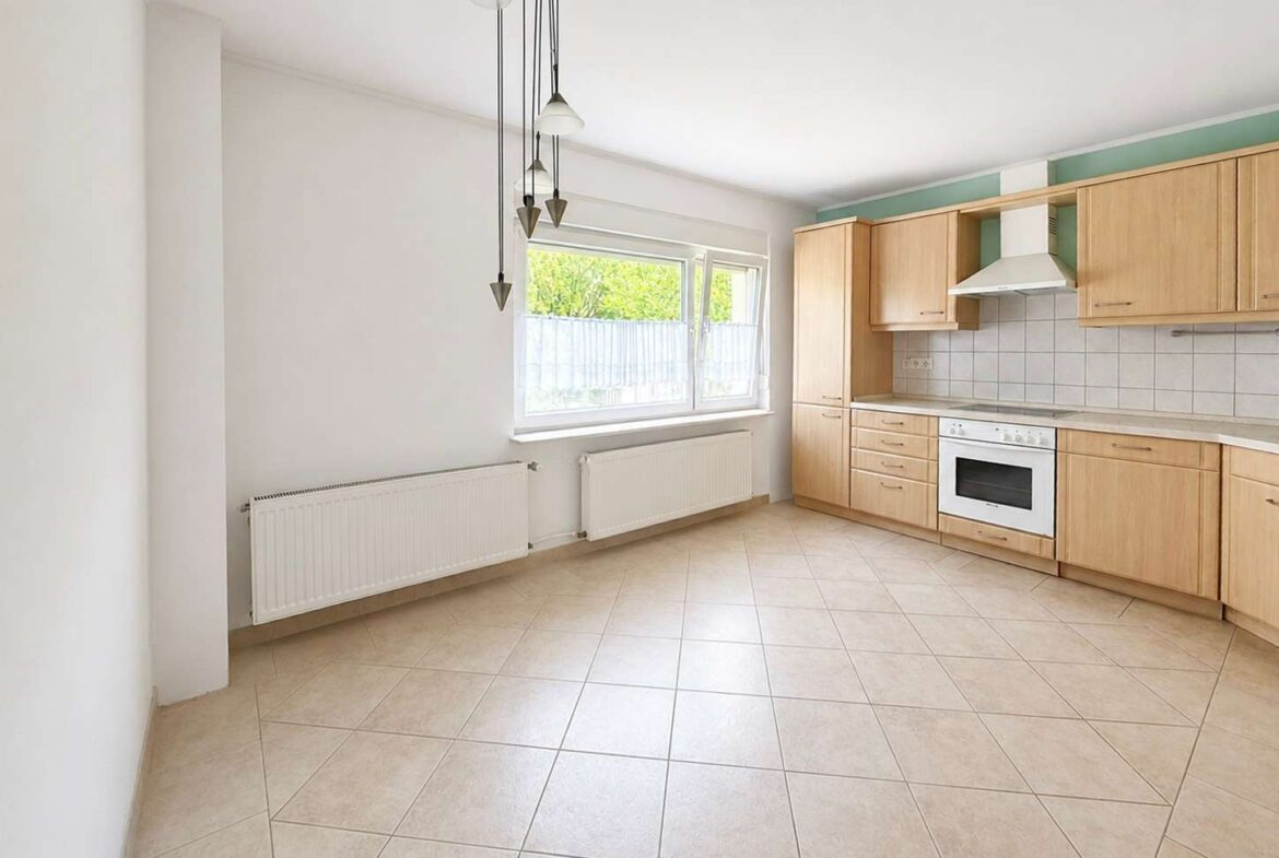 Bright L-shaped kitchen with light wood cabinets, built-in oven, range hood, tiled backsplash, and a large window.