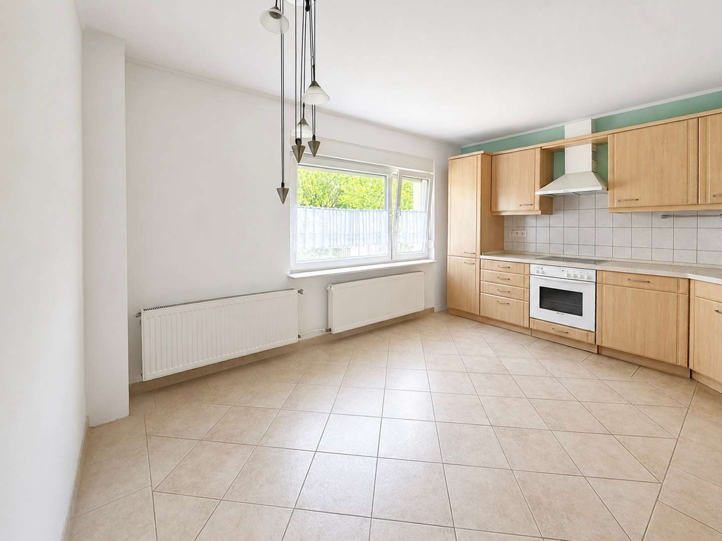 Bright L-shaped kitchen with light wood cabinets, built-in oven, range hood, tiled backsplash, and a large window.