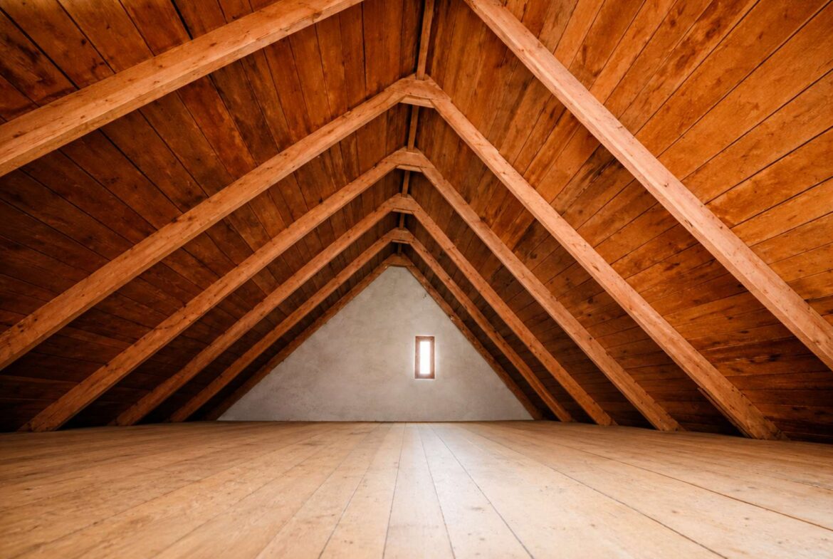 A wide attic space with exposed wooden beams forming triangles, empty floor, and a small vertical window at the far wall.