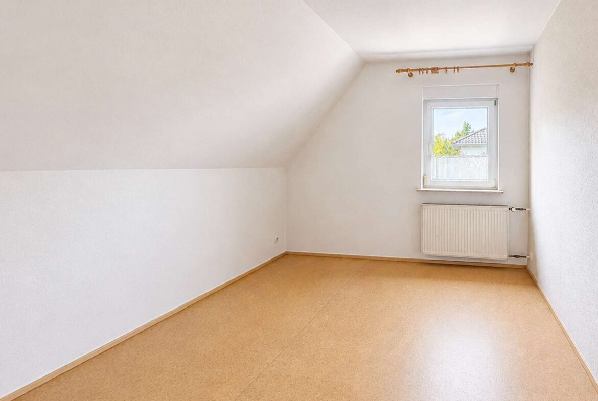 Empty white attic-like room with a sloped ceiling on the left, a small window above a radiator, and beige flooring.