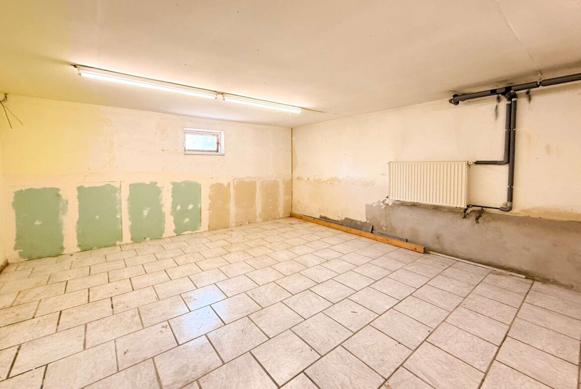 A bare, unfinished basement room with white tiled floor, pale walls with green patches, a long fluorescent light fixture on the ceiling, and exposed pipes along the right wall beneath a radiator.