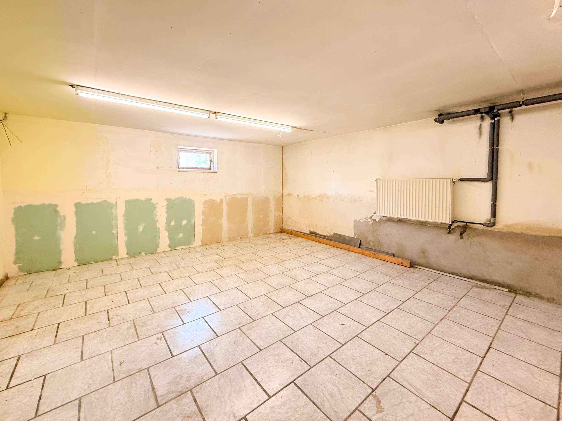 A bare, unfinished basement room with white tiled floor, pale walls with green patches, a long fluorescent light fixture on the ceiling, and exposed pipes along the right wall beneath a radiator.
