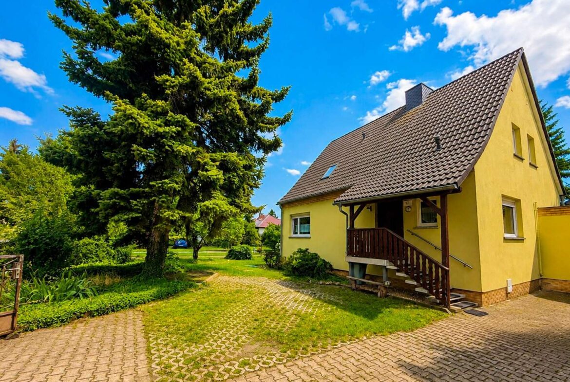 Yellow house with brown tiled roof and wooden porch, beside a large evergreen tree on a sunny day.