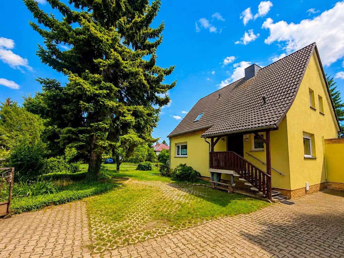 Yellow house with brown tiled roof and wooden porch, beside a large evergreen tree on a sunny day.