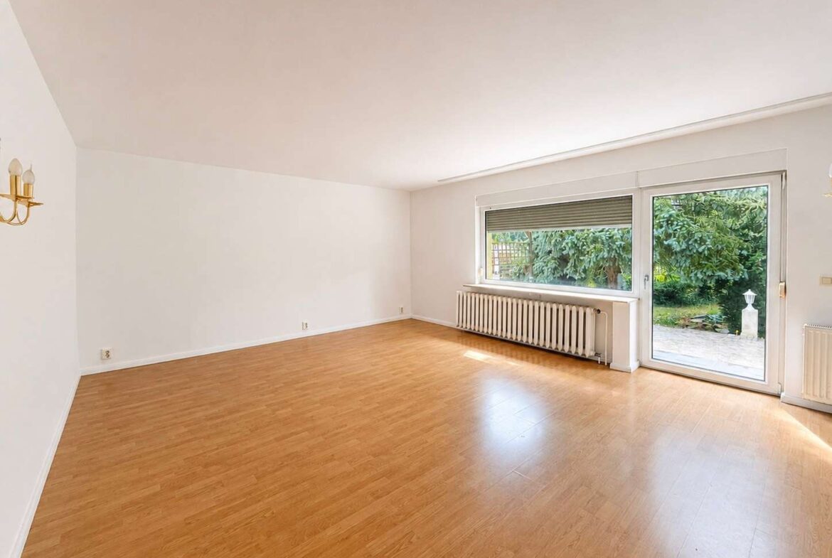 Empty living room with light wood floor, white walls, and large glass door opening to a green garden patio area; radiator along wall under window.