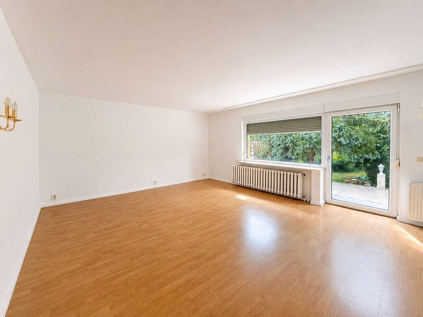 Empty living room with light wood floor, white walls, and large glass door opening to a green garden patio area; radiator along wall under window.