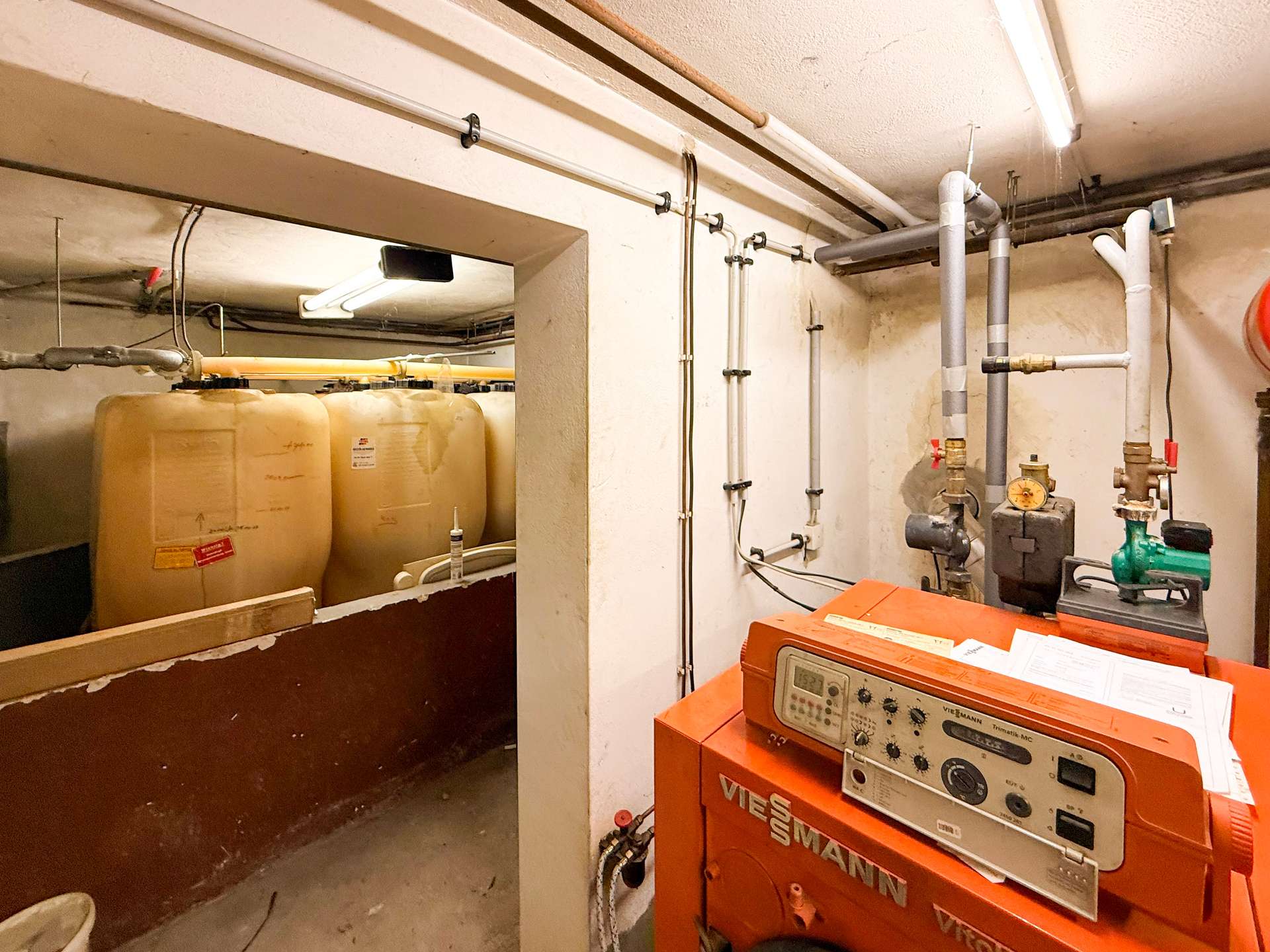 Utility room with an orange Viessmann boiler and large white water storage tanks connected by pipes along the walls, in a dim basement area.